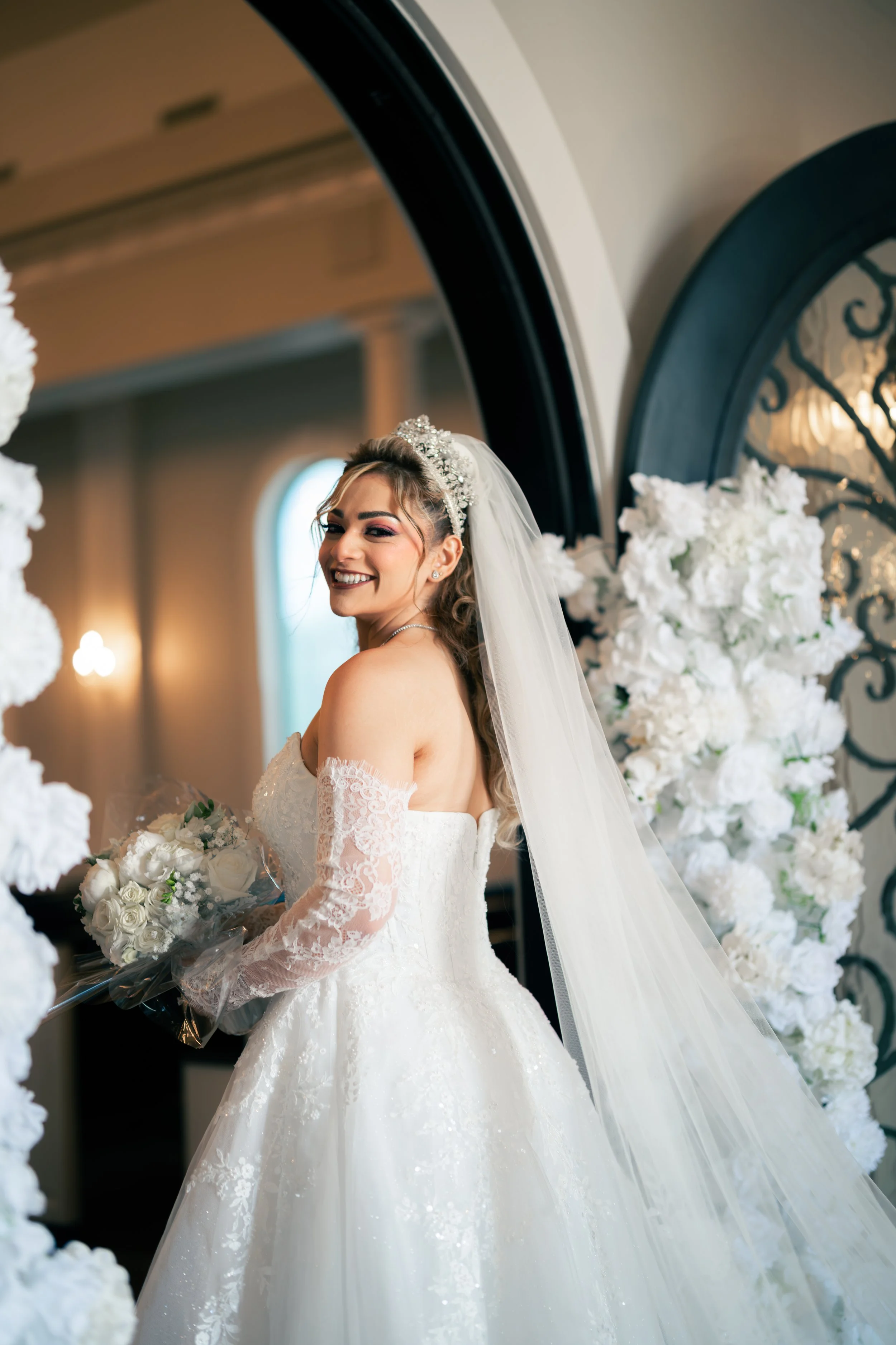 A bride in a white wedding dress with lace sleeves, smiling while holding a bouquet of white roses, standing near a floral arch with white flowers and a decorative mirror behind her.