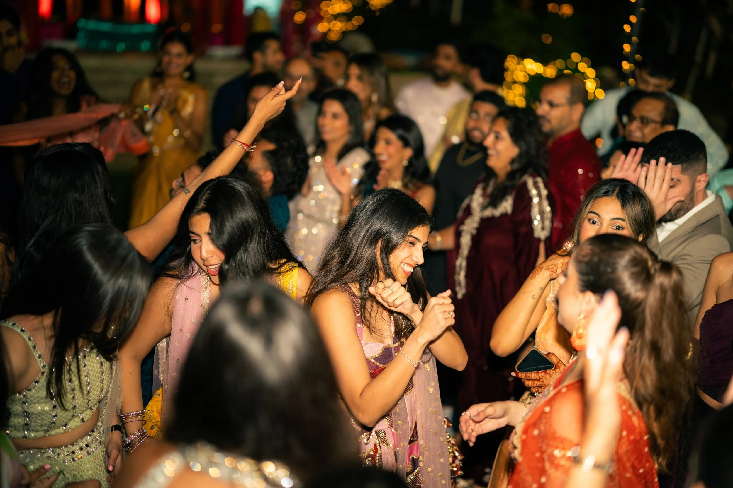 People dancing and celebrating at a lively indoor party with colorful traditional attire, bright lighting, and festive decorations in the background.