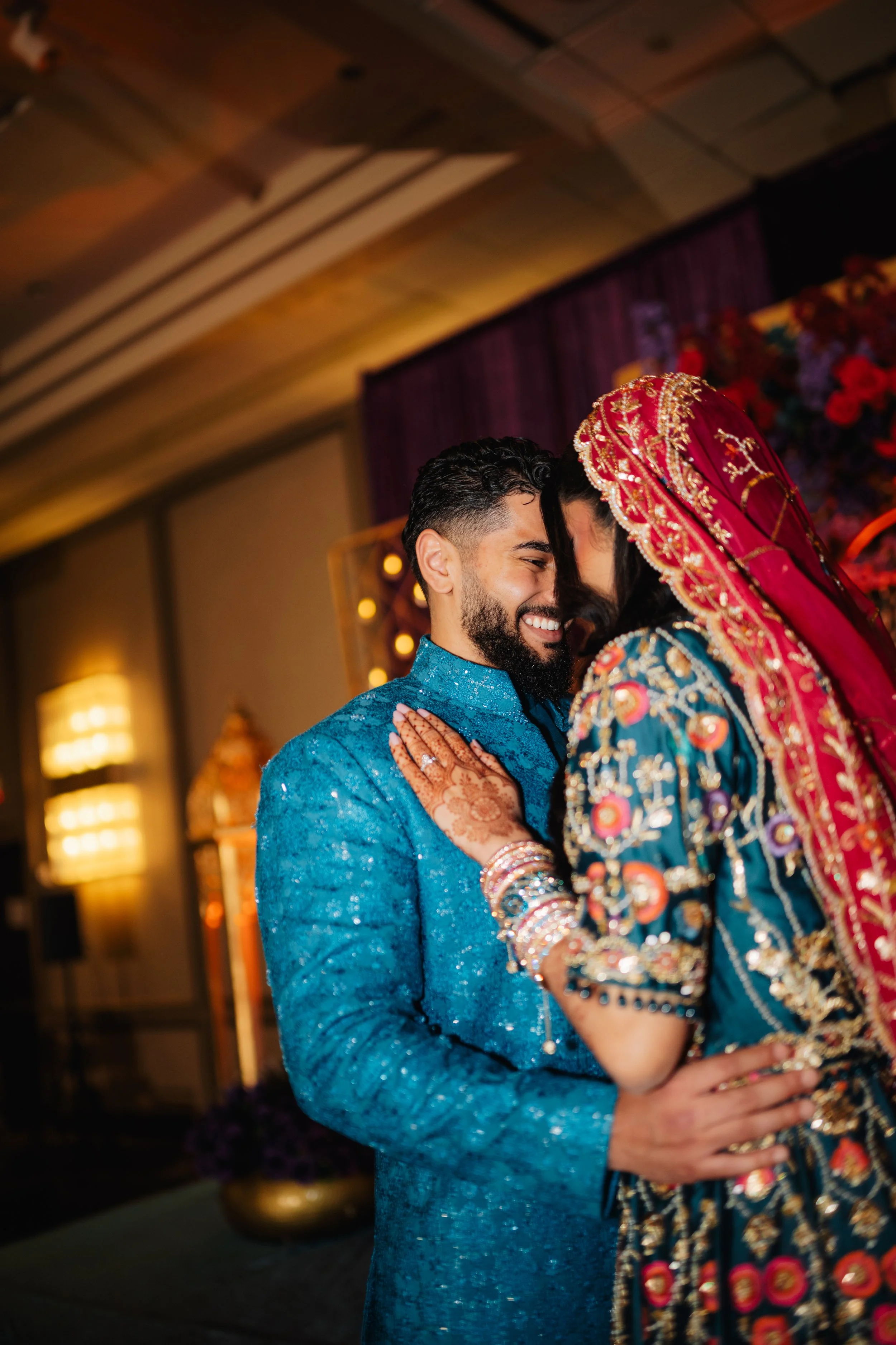 A couple dressed in traditional Indian wedding attire sharing a close moment, with the bride in a richly embroidered red and gold dupatta covering her head and the groom in a blue sherwani, both smiling with their foreheads touching.