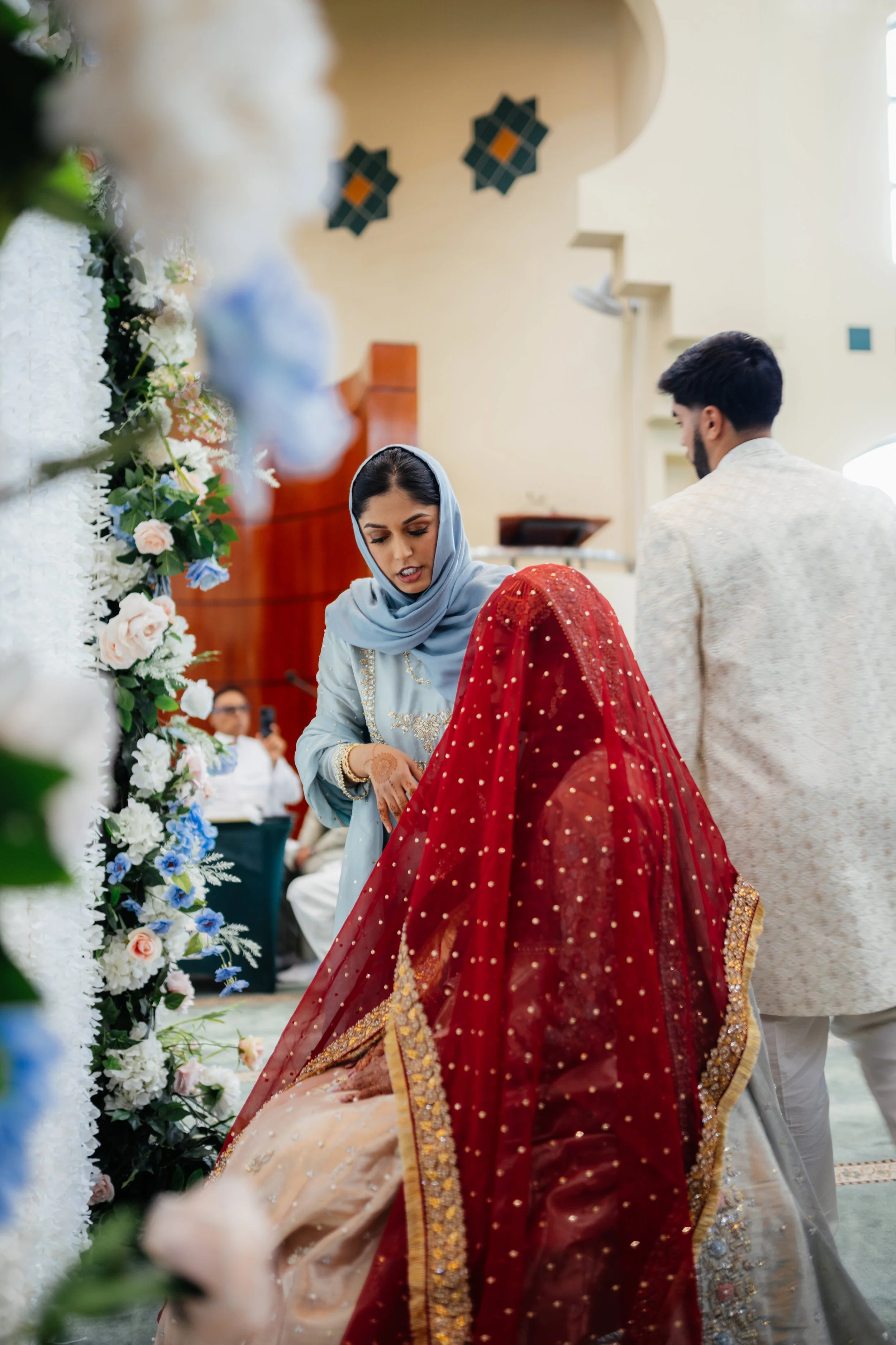 A woman in traditional South Asian wedding attire kneels on the floor, wearing a richly embroidered red and gold bridal dress, with a man in a cream-colored outfit standing nearby. They are under a floral wedding arch at a decorated indoor venue.