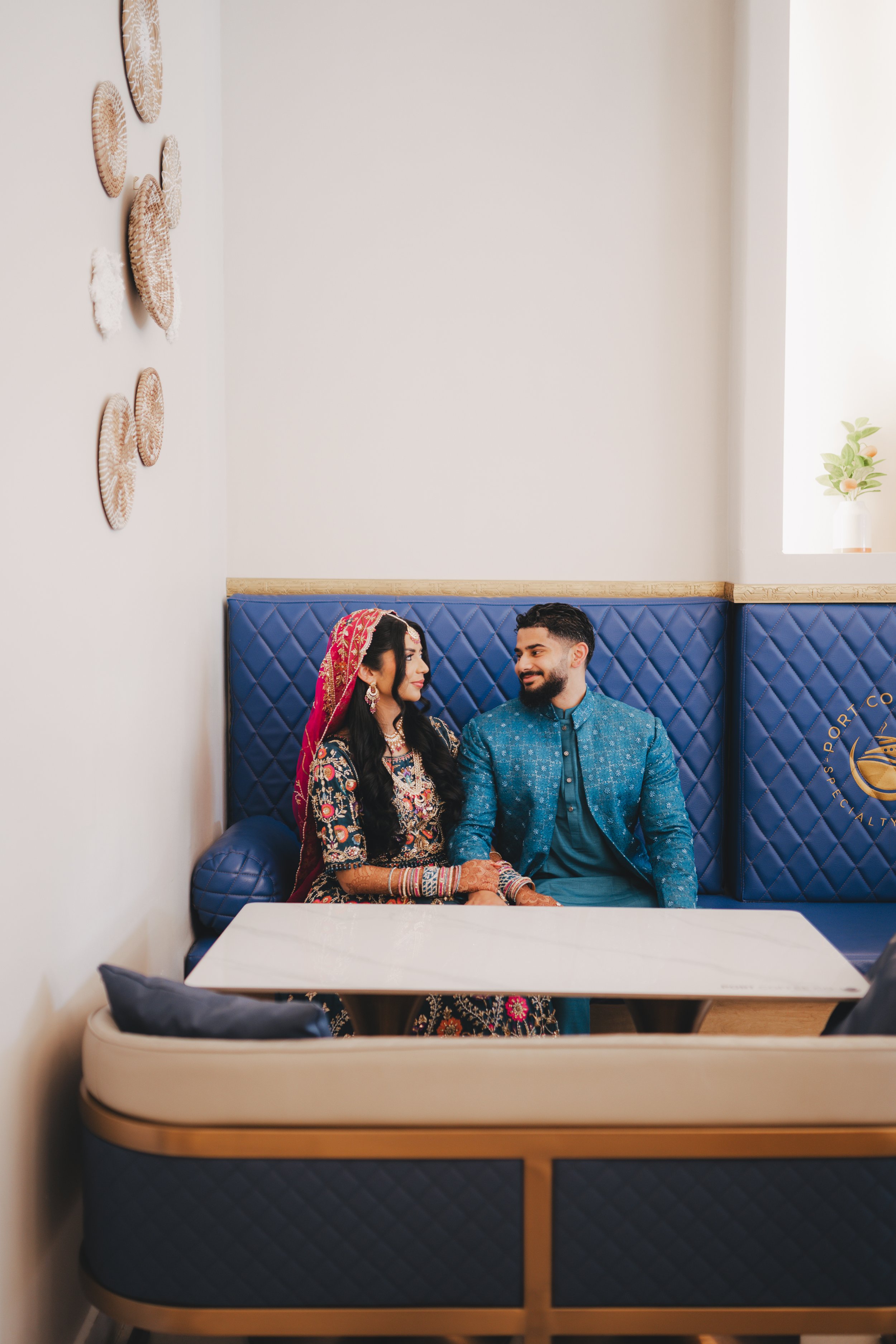 A newlywed Indian couple in traditional attire sitting on a blue quilted booth in a modern restaurant, engaged in a conversation, with a white table in front of them and beige decor with wall art on the side wall.