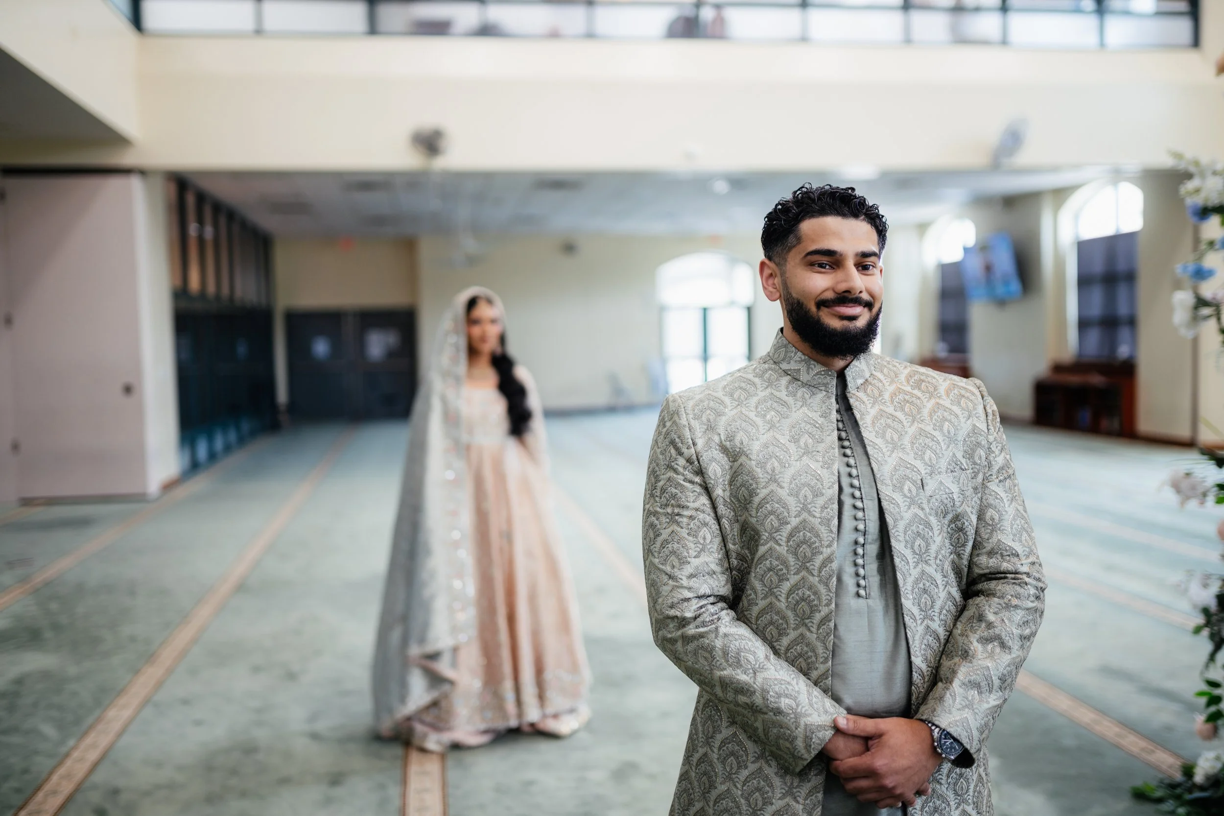 A man in traditional Indian attire smiling with his hands clasped in front of him, standing in a decorated indoor venue with a woman in traditional dress in the background.