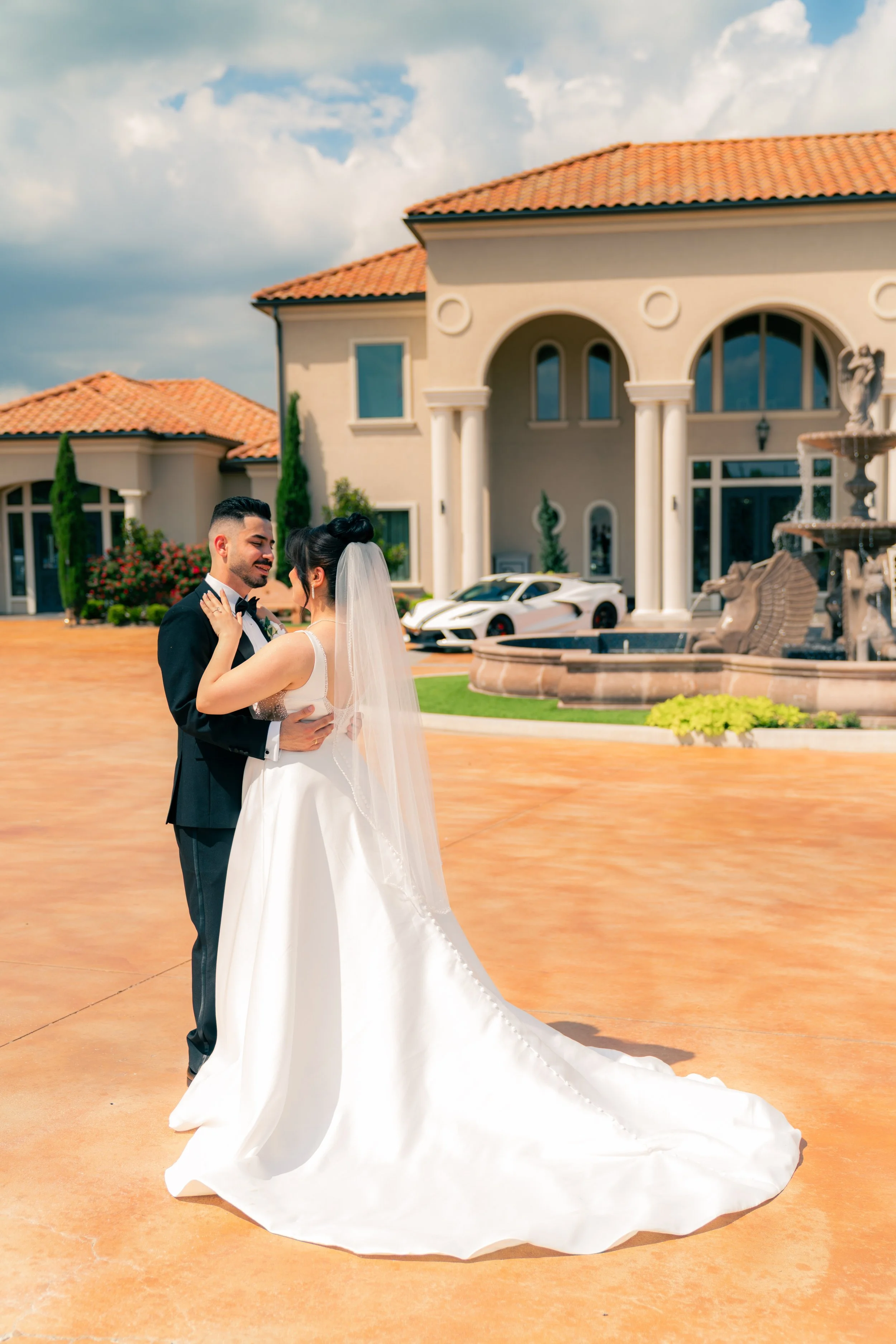 A newlywed couple dancing outside a large, elegant house with a fountain and luxury cars in the driveway.