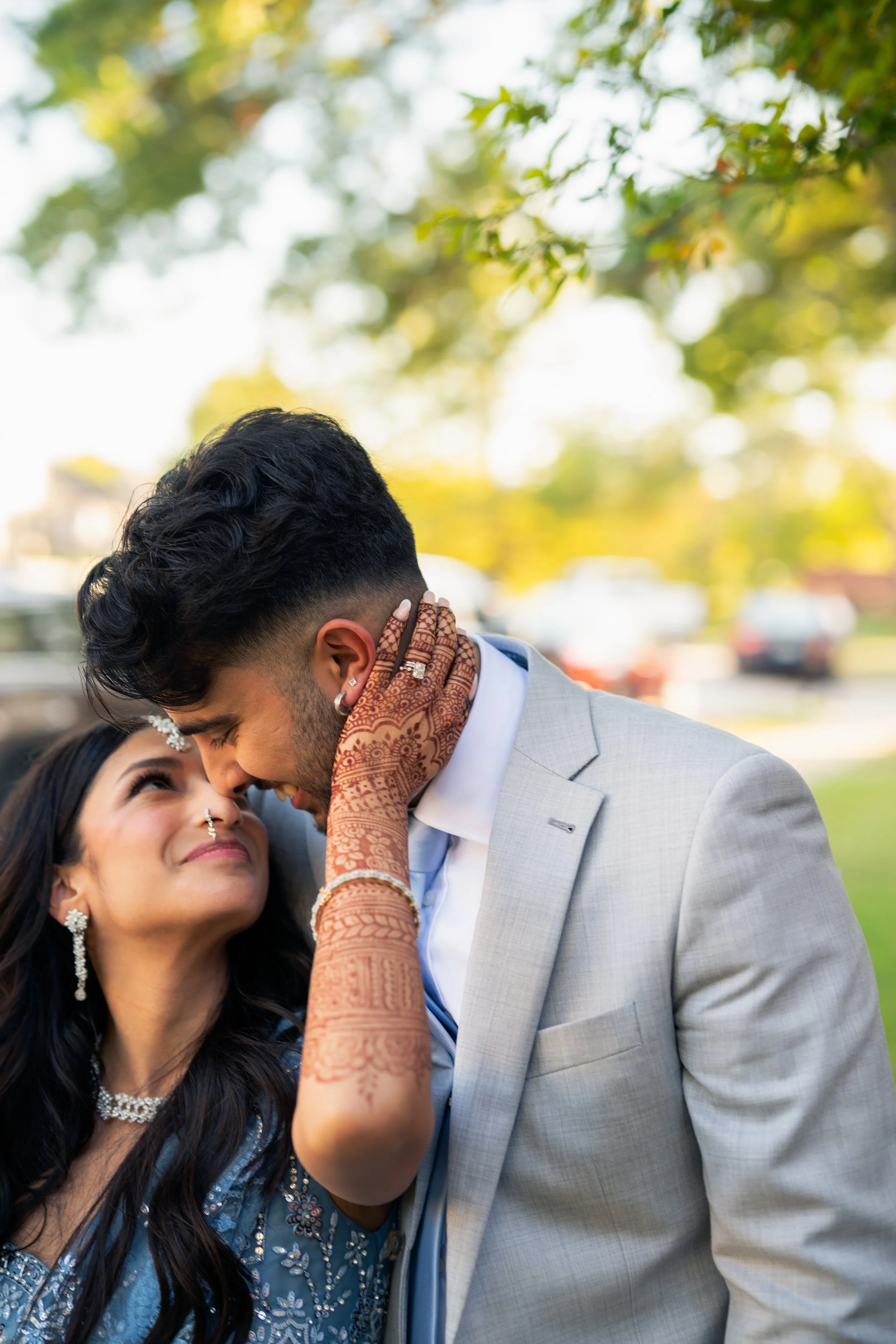 A couple of Indian ethnicity embracing outdoors during daytime, with the woman smiling and the man leaning in, dressed in formal attire.