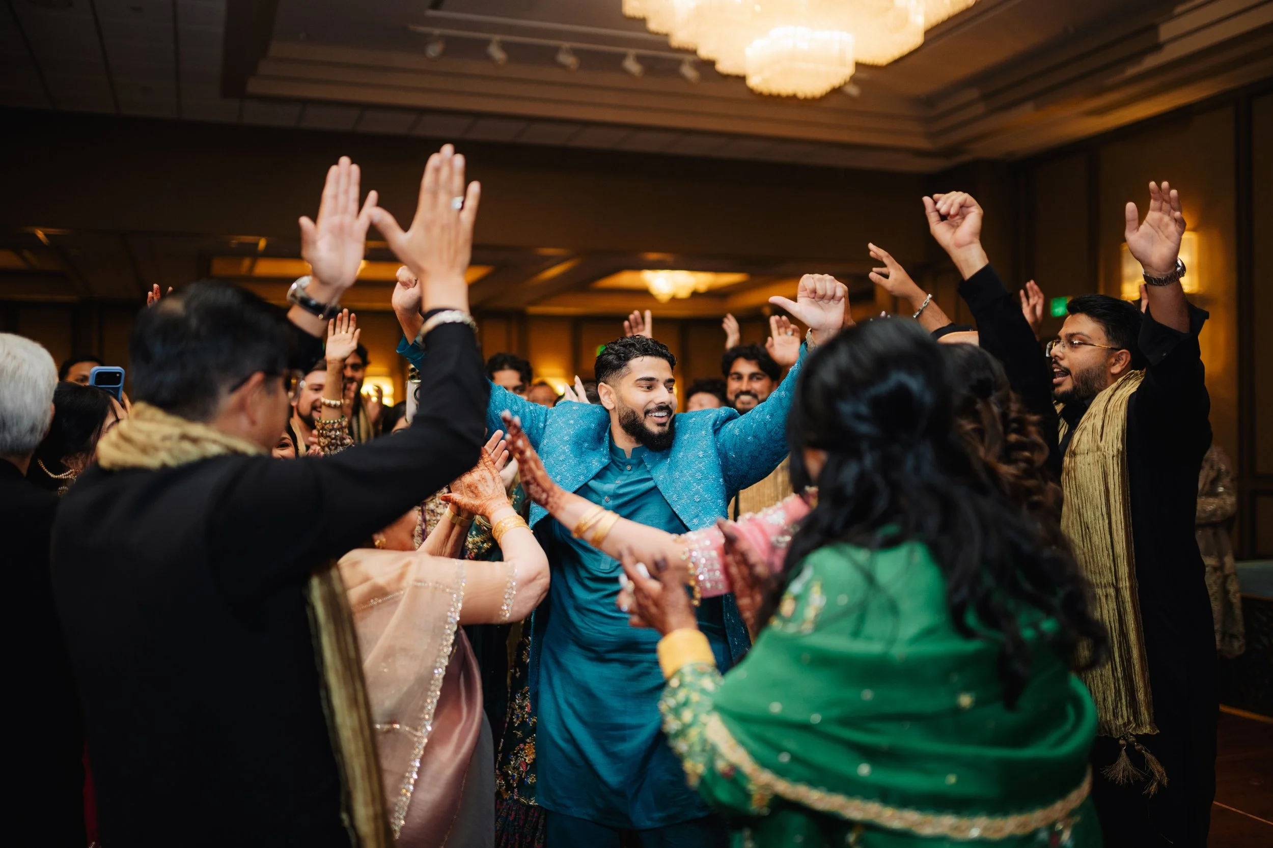 People celebrating at an indoor wedding or festive event, dancing and smiling, with a chandelier overhead and warm lighting in the background.