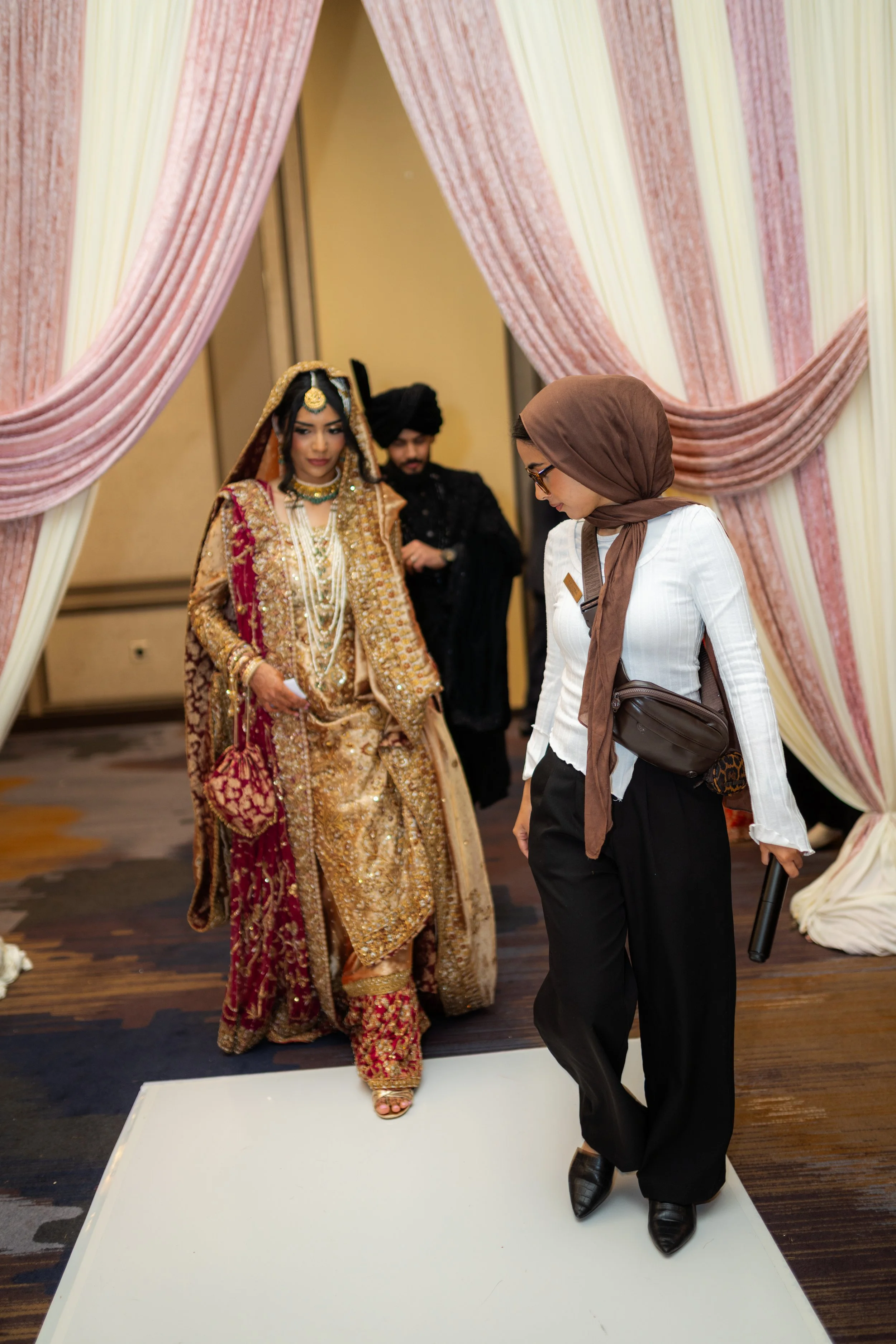 A woman dressed in traditional Indian wedding attire, standing on a white platform, surrounded by pink drapes, with a person in modern attire standing nearby.
