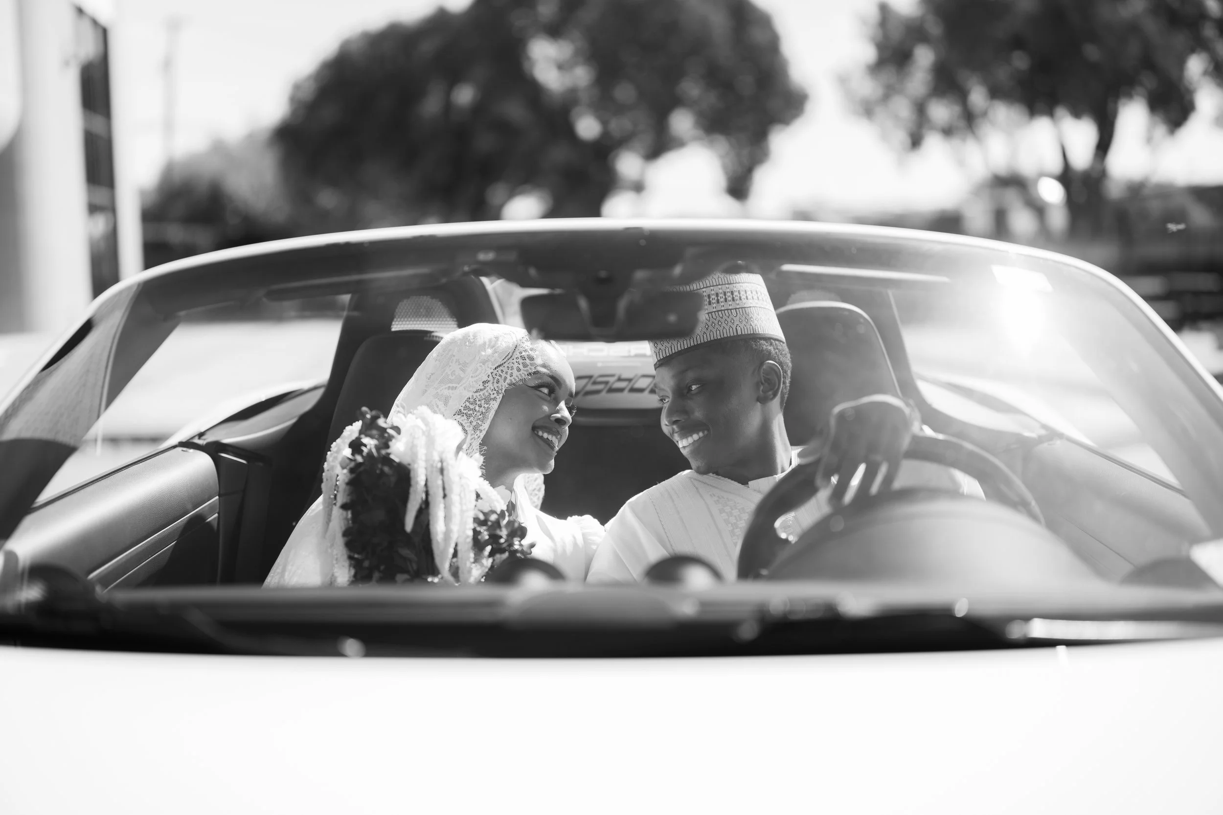 A black and white photo of a bride and groom sitting in a car, smiling at each other, with the bride wearing a lace headscarf and holding flowers, and the groom wearing a traditional cap and kurta.