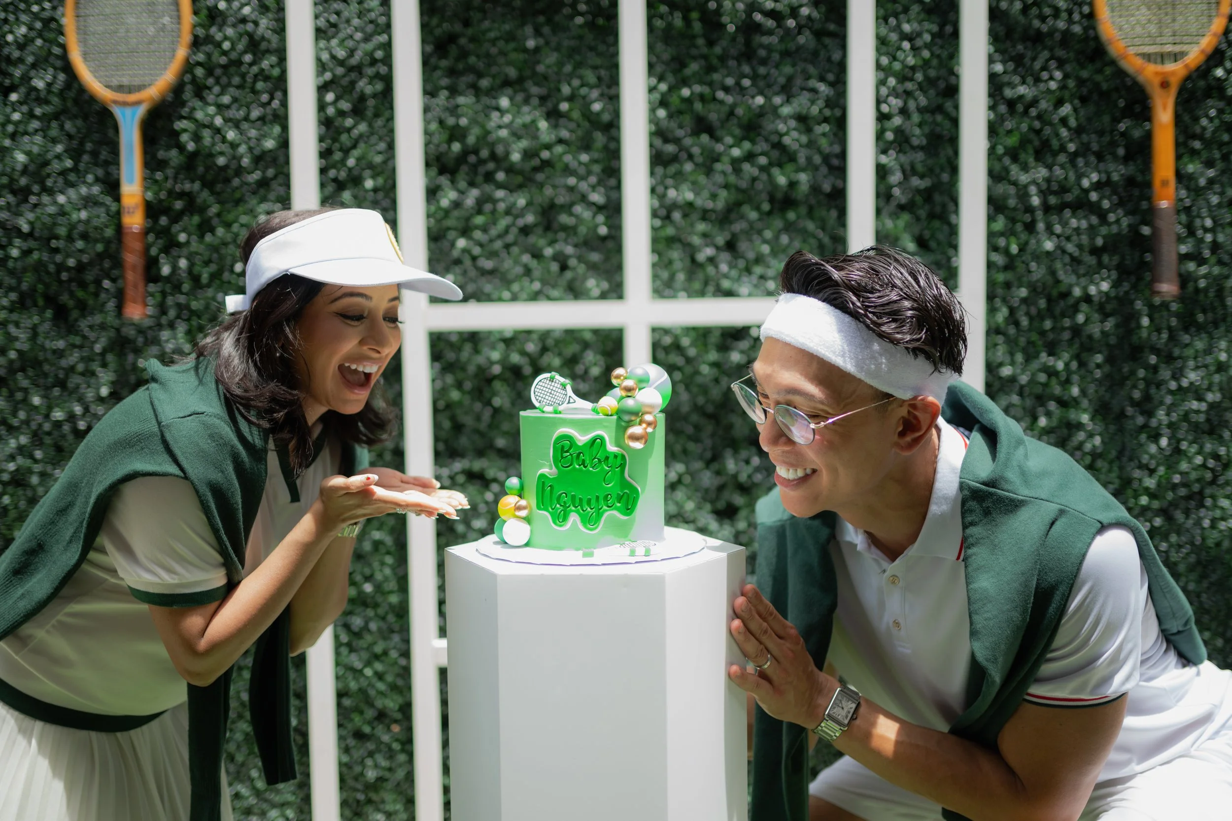 Two people are celebrating a birthday outdoors in front of a green hedge, wearing green and white clothing and white headbands. They are blowing out candles on a small, bright green birthday cake decorated with colorful spheres and a miniature tennis