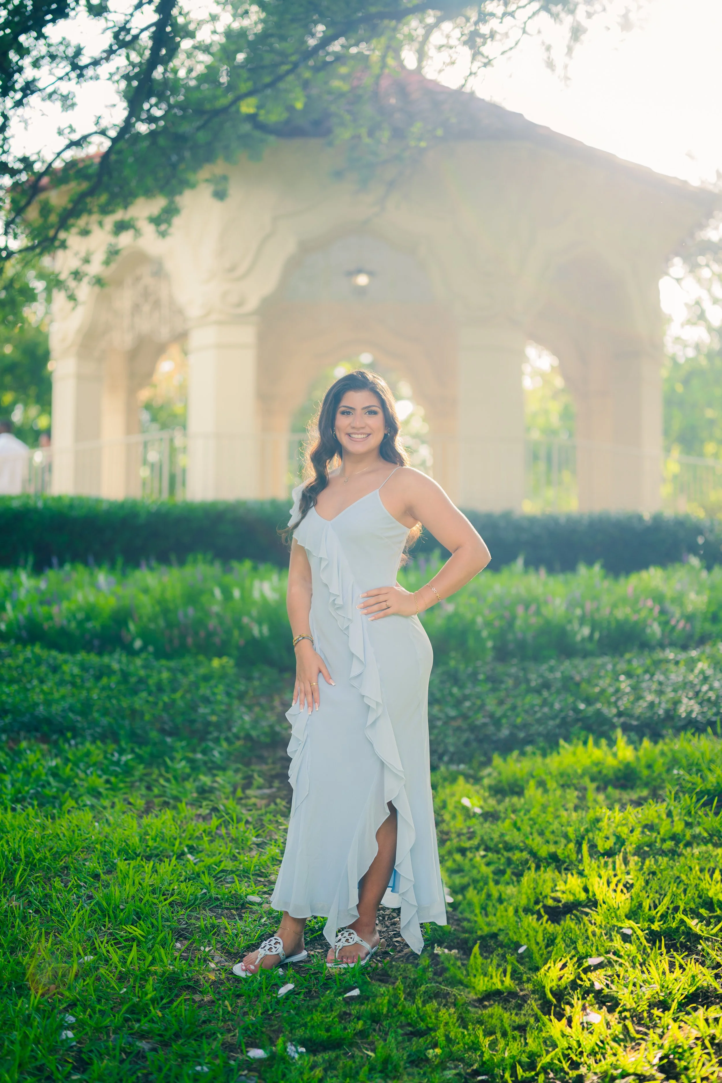 A woman in a white dress standing on grass in front of a decorative pavilion, with sunlight filtering through trees behind her.