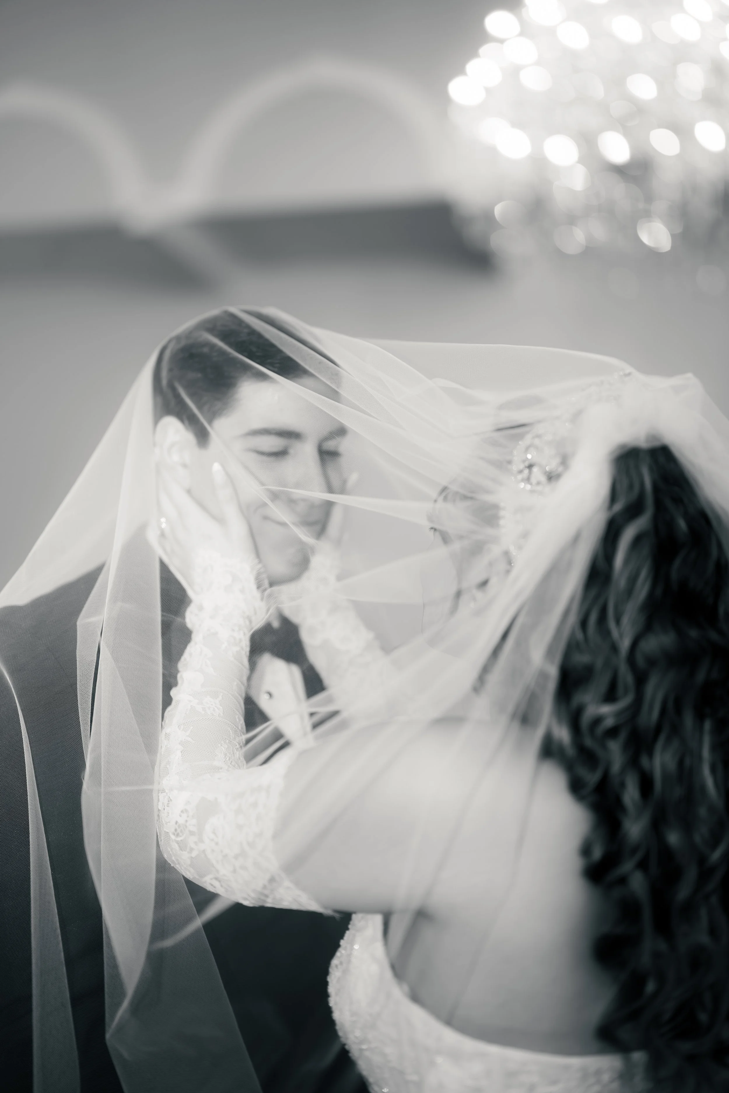 A bride and groom share a moment behind the bride's veil at their wedding, with the bride smiling and touching the groom's face.