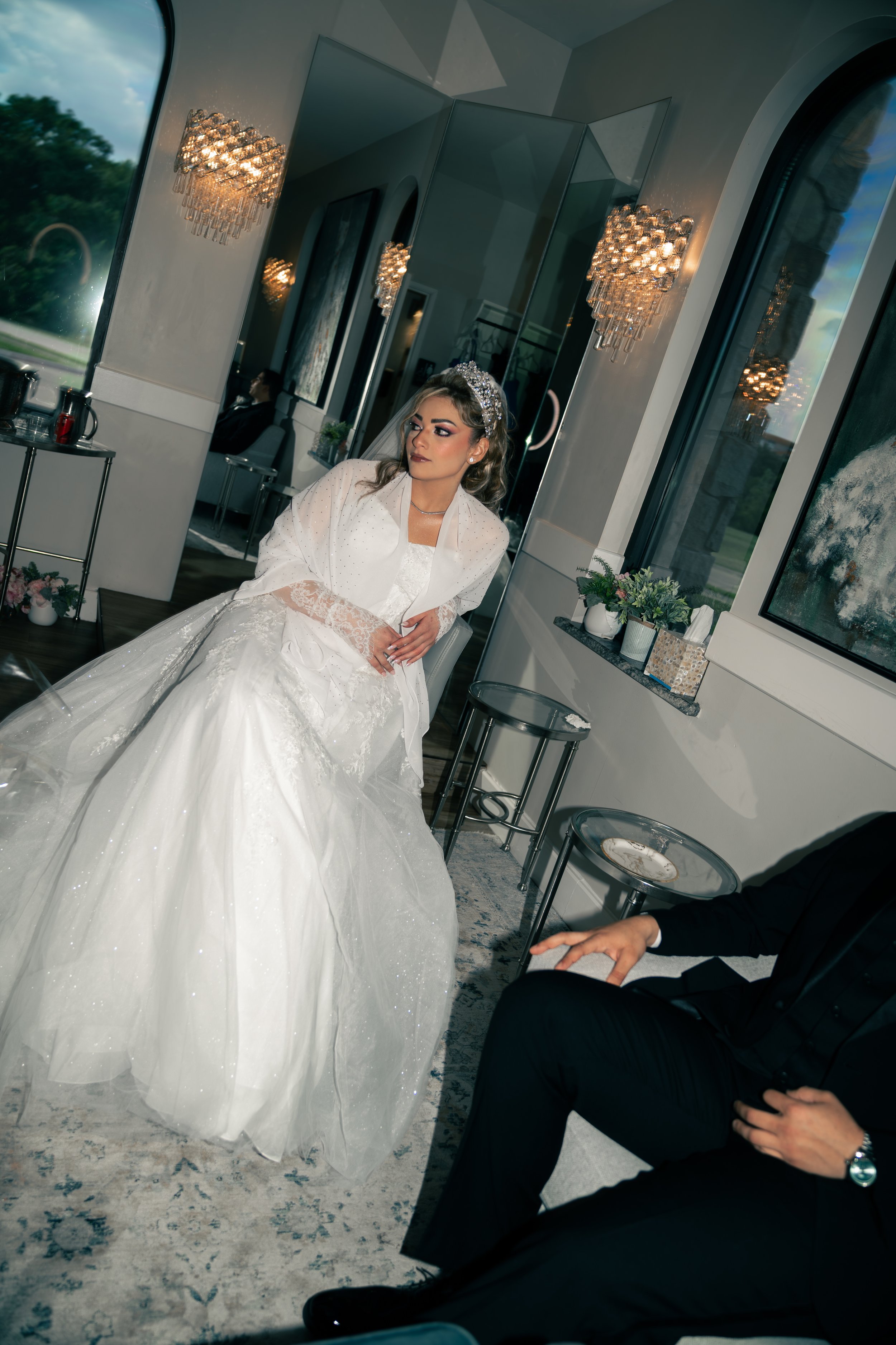 A bride in a white wedding gown and veil sitting on a chair indoors, with a groom dressed in a black suit sitting nearby. The room has large windows, modern decor, and elegant lighting fixtures.