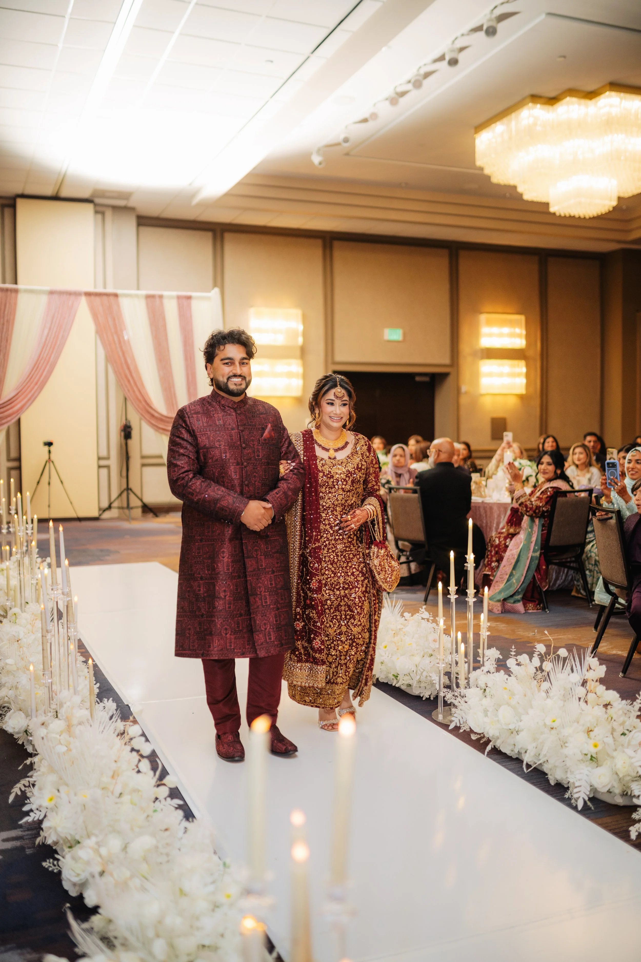 A couple dressed in traditional Indian wedding attire walking down a white aisle lined with white flowers and candles at a wedding reception. The man is wearing a maroon sherwani, and the woman is wearing a maroon and gold lehenga. Guests are seated 