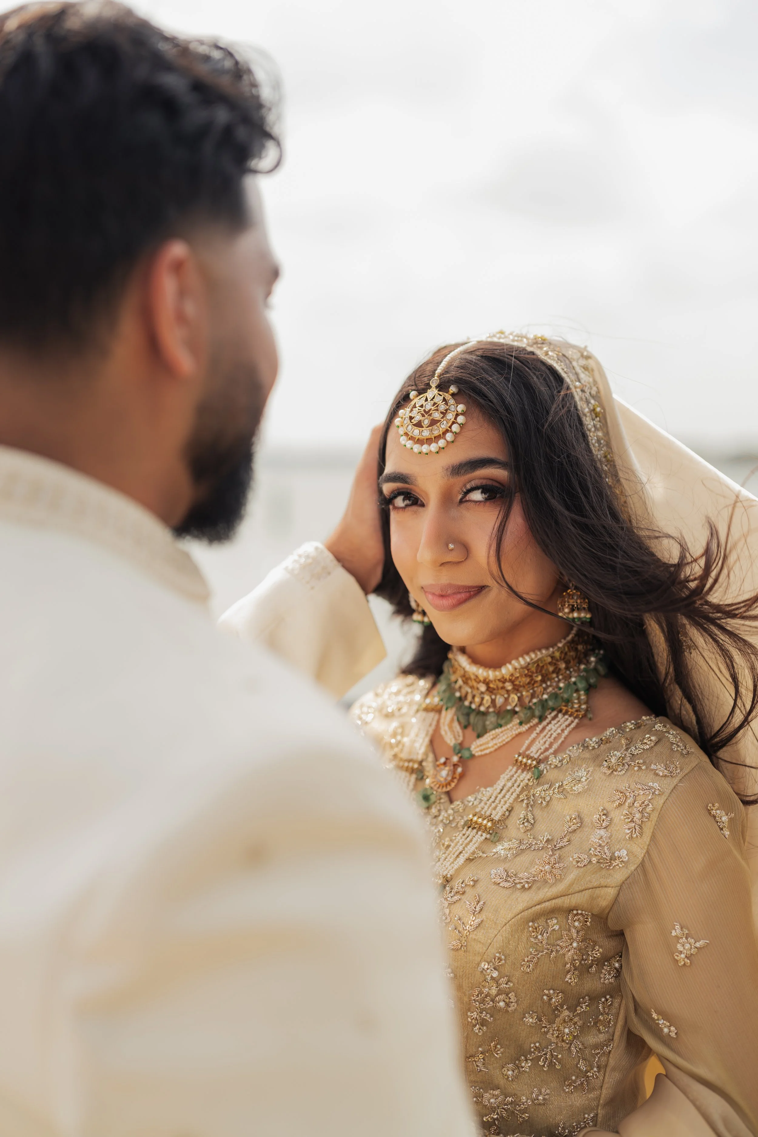A woman dressed in traditional Indian bridal attire, adorned with intricate gold jewelry including a maang tikka, nose ring, earrings, necklace, and embroidered beige fabric with floral patterns. A man, seen from the back, appears to be wearing a cre