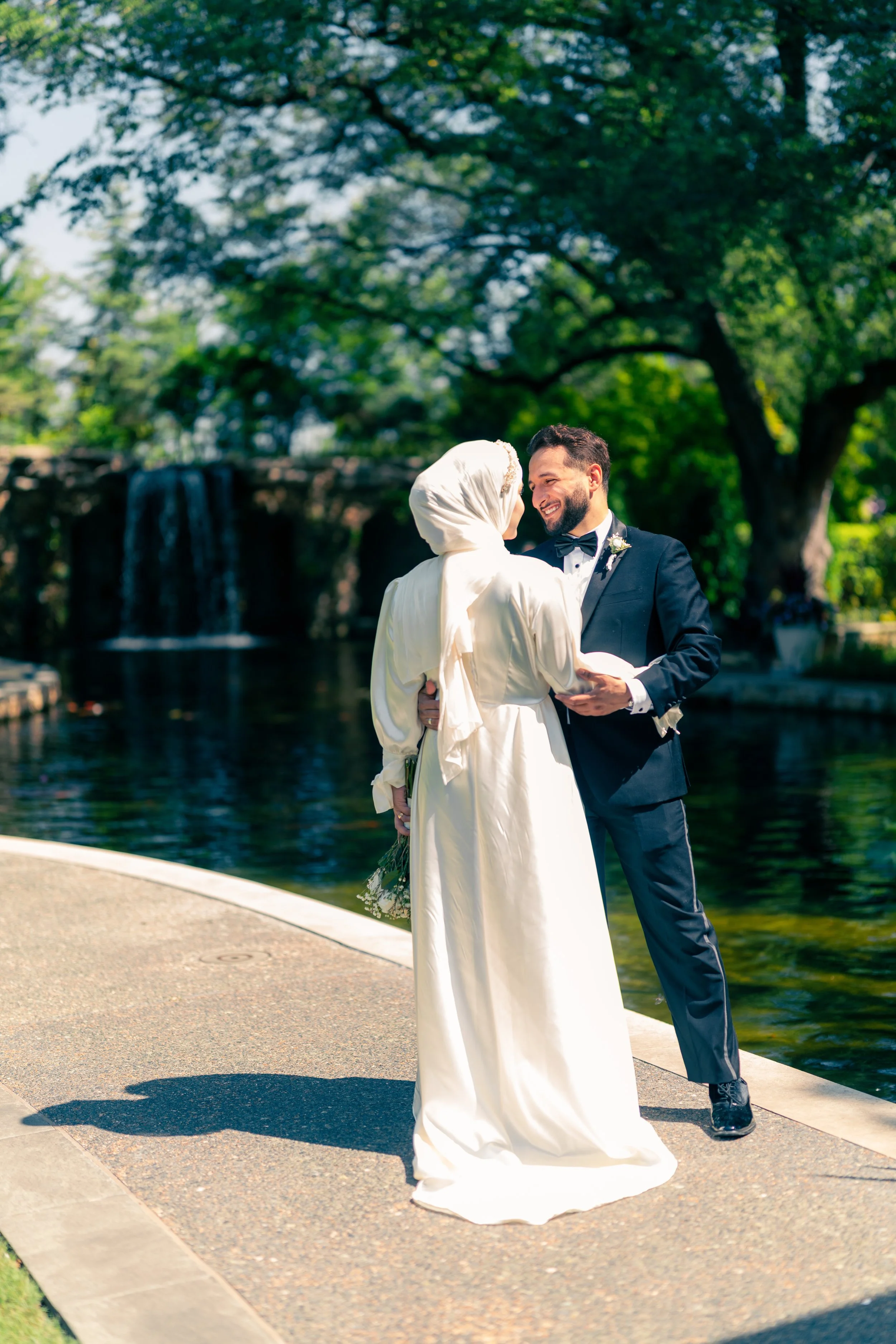 A bride in a white dress and headscarf and a groom in a tuxedo are smiling and holding hands by a pond with a waterfall in the background.