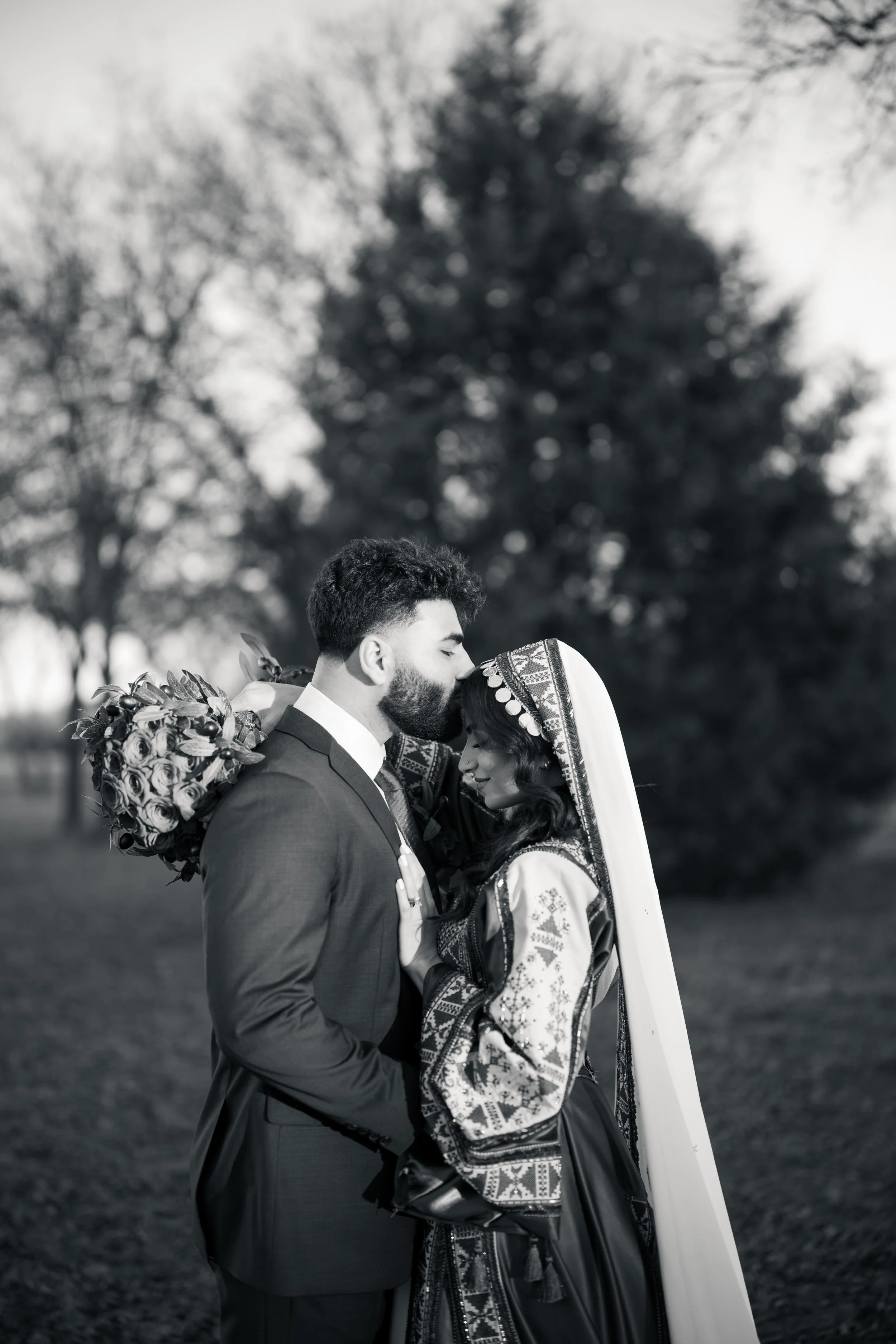 A black-and-white photo of a couple on their wedding day, with the groom kissing the forehead of the bride, who is wearing traditional cultural attire. The groom holds a bouquet of roses over his shoulder. They stand outdoors with trees in the backgr