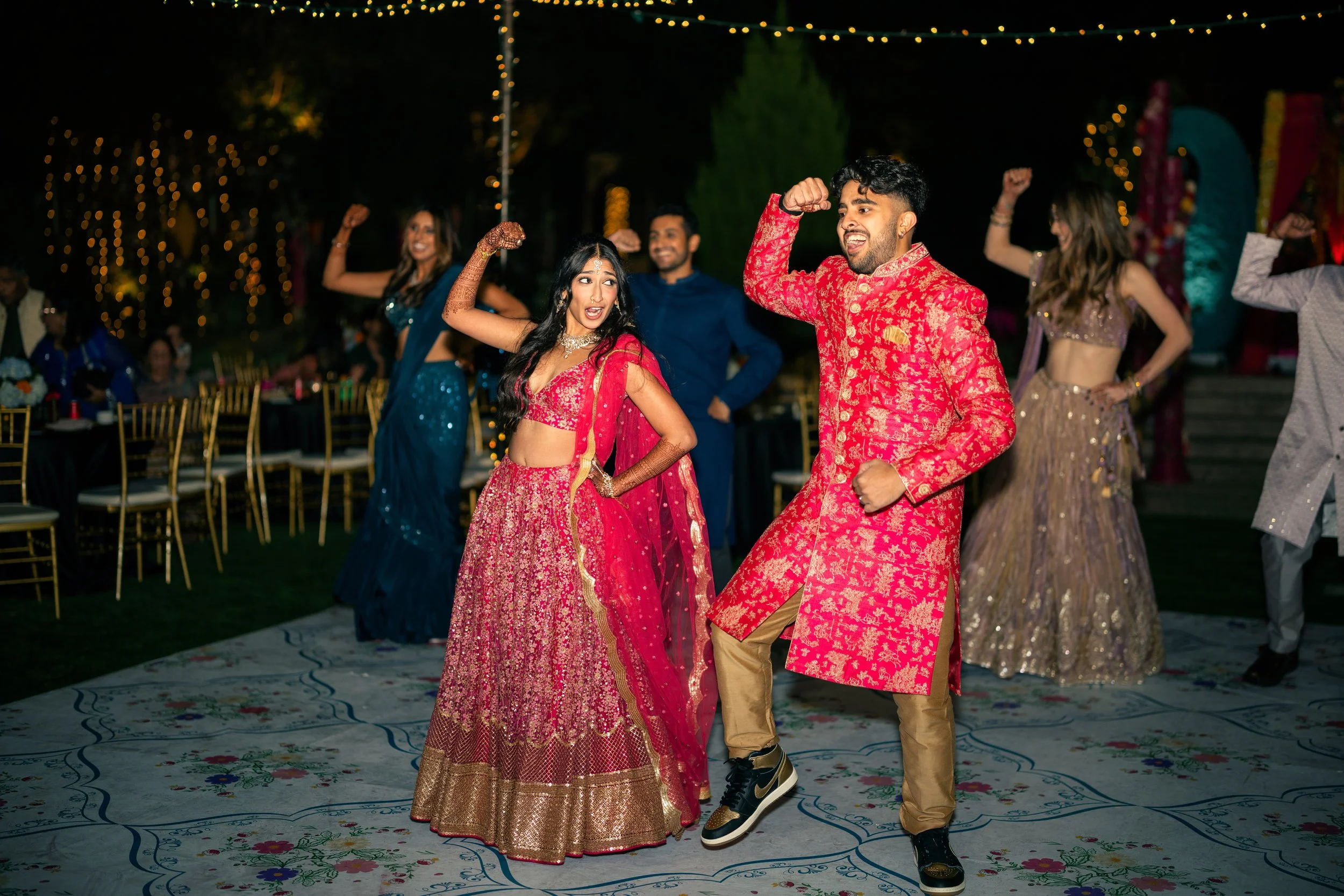 People dancing at a wedding reception, with women in colorful sarees and women in embellished lehengas, and men in traditional kurtas, on a decorated outdoor dance floor at night.