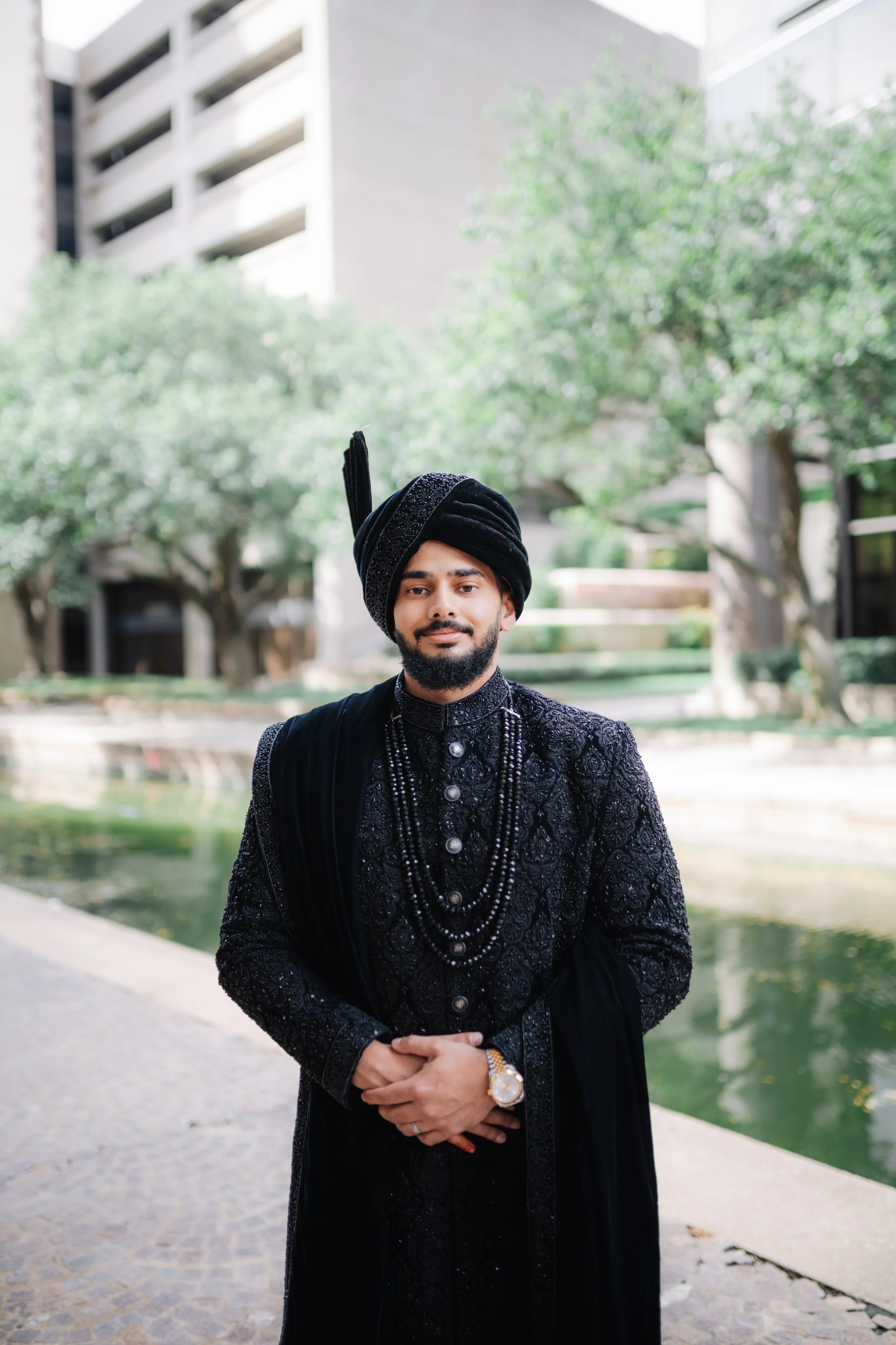 A man dressed in traditional South Asian wedding attire, including a black embroidered sherwani, black turban with a feather, and layered necklaces, standing outdoors by a small water feature with trees and a modern building in the background.