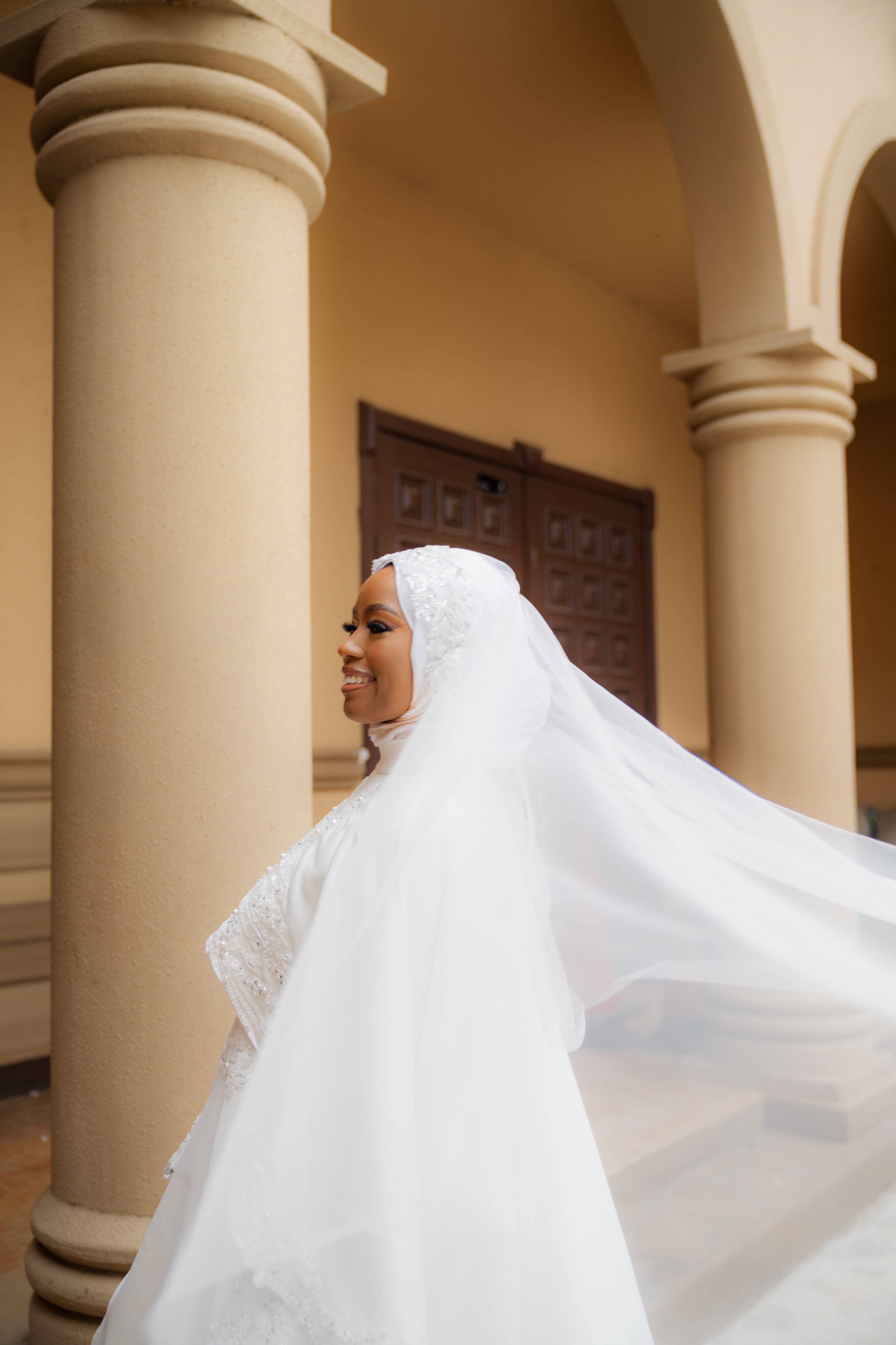 A bride in a white wedding dress and veil standing in front of beige columns and arches, smiling.