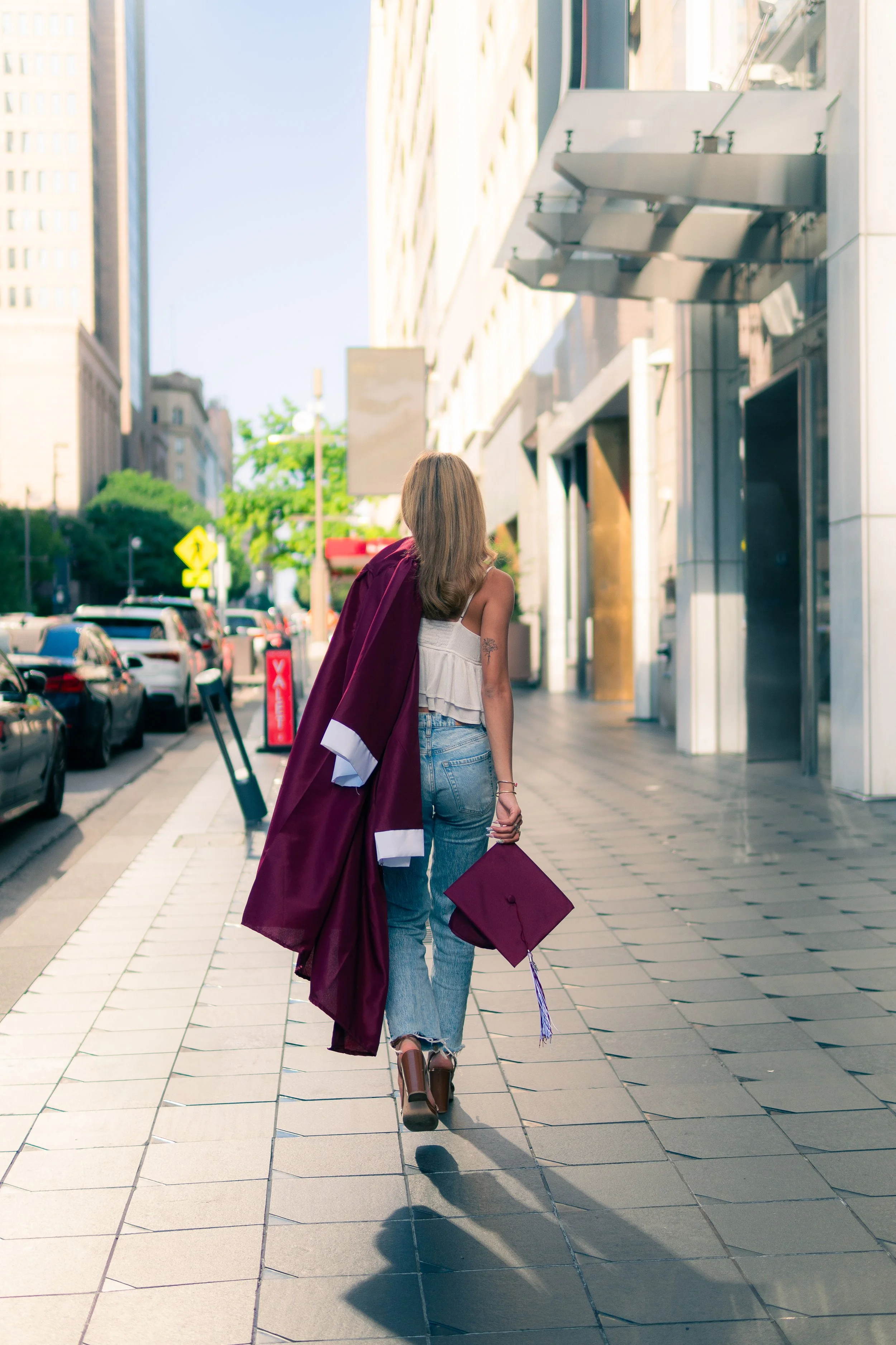A woman in casual clothes, carrying a graduation cap and gown, walking on a city sidewalk during daytime.