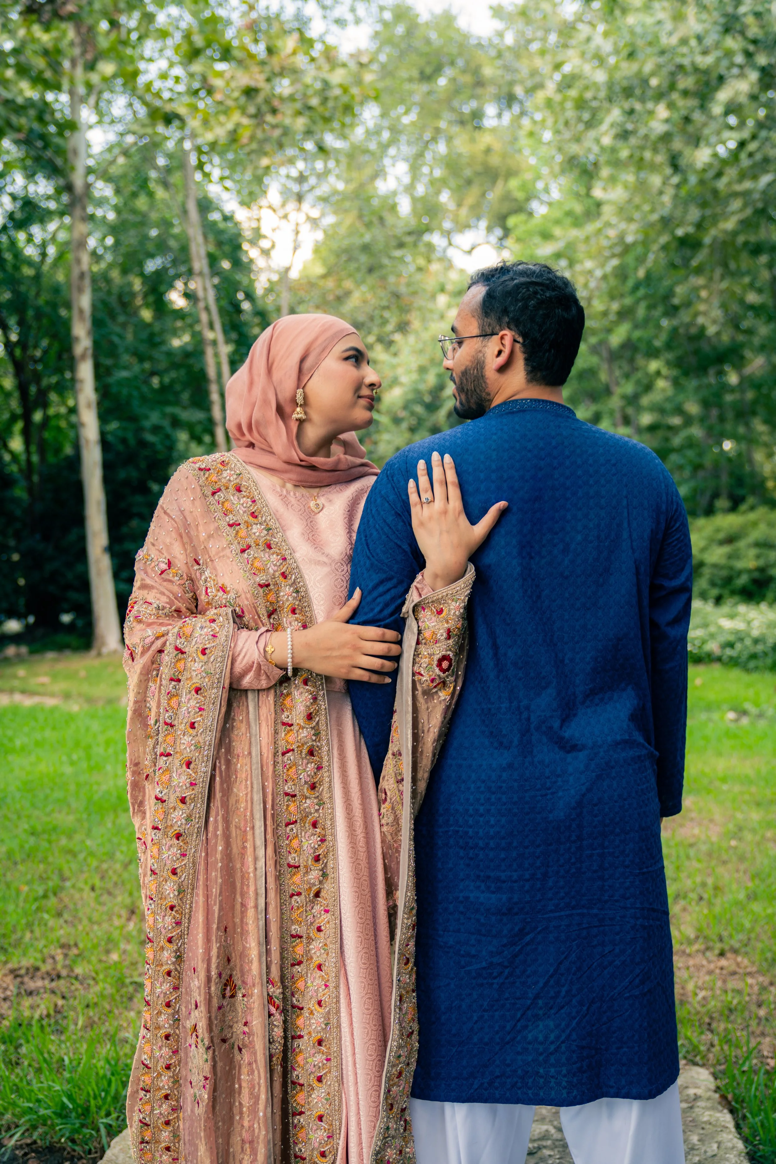 A couple dressed in traditional South Asian attire standing close together in a park with green trees and grass.