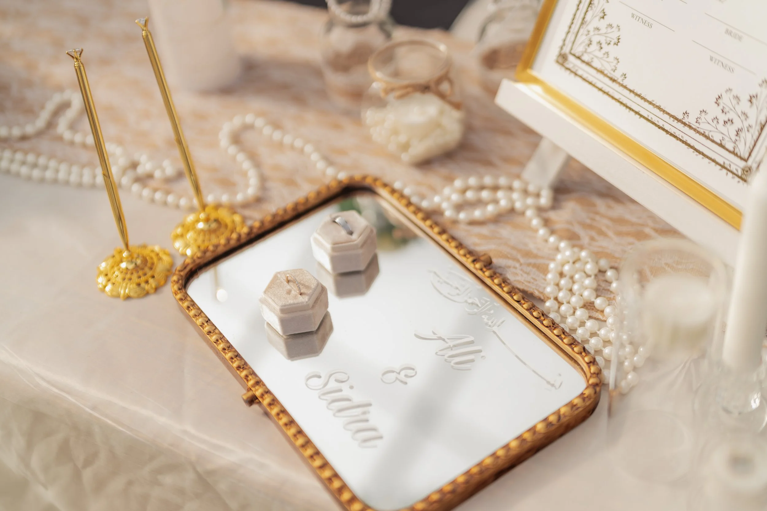 A jewelry display tray with pearl necklaces, gold and pearl earrings, pearl bracelet, and two small jewelry boxes, along with a partially visible book or picture frame with gold edges in the background.