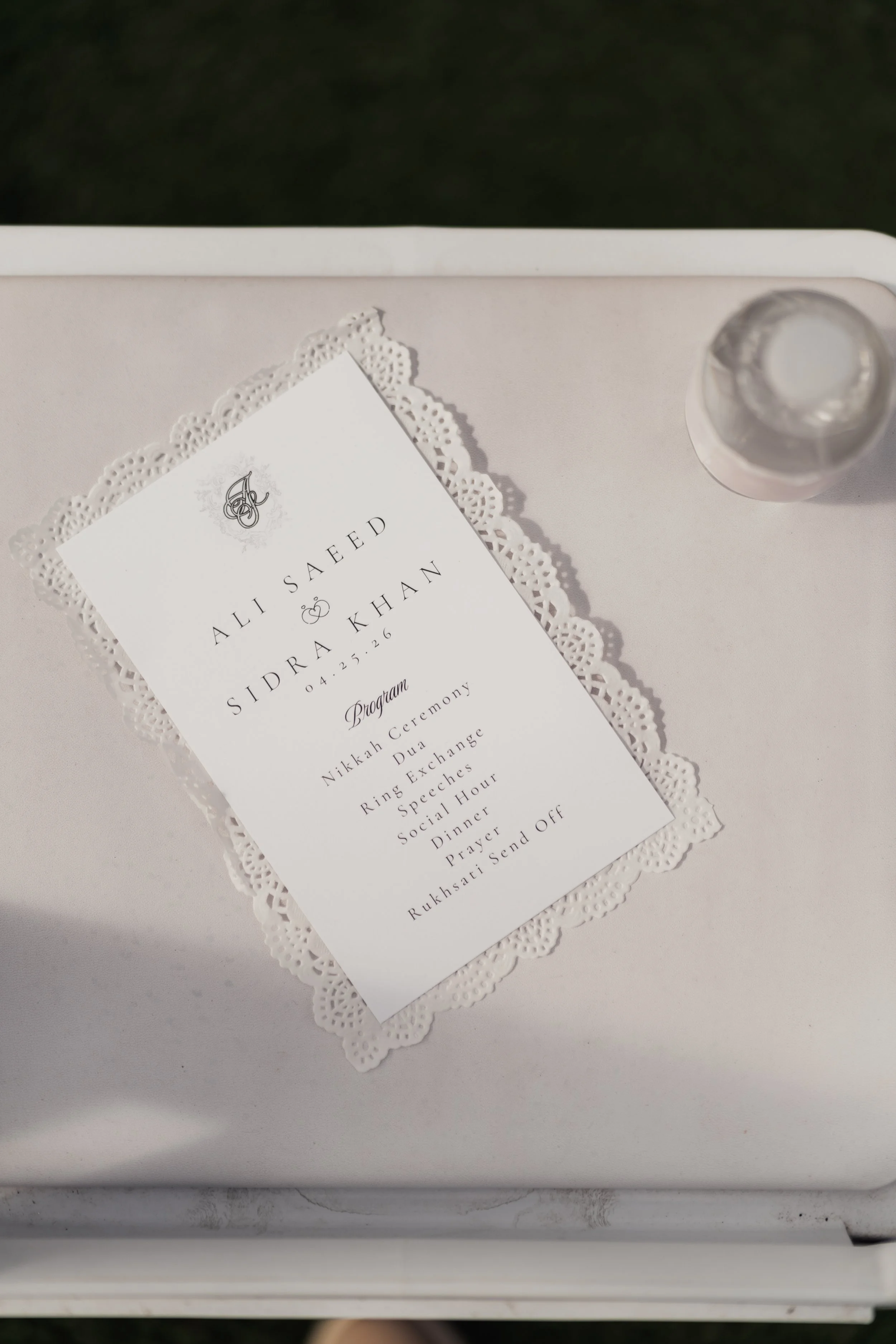 A wedding invitation card on a white doily, placed on a white surface, with a small silver bottle to the right.