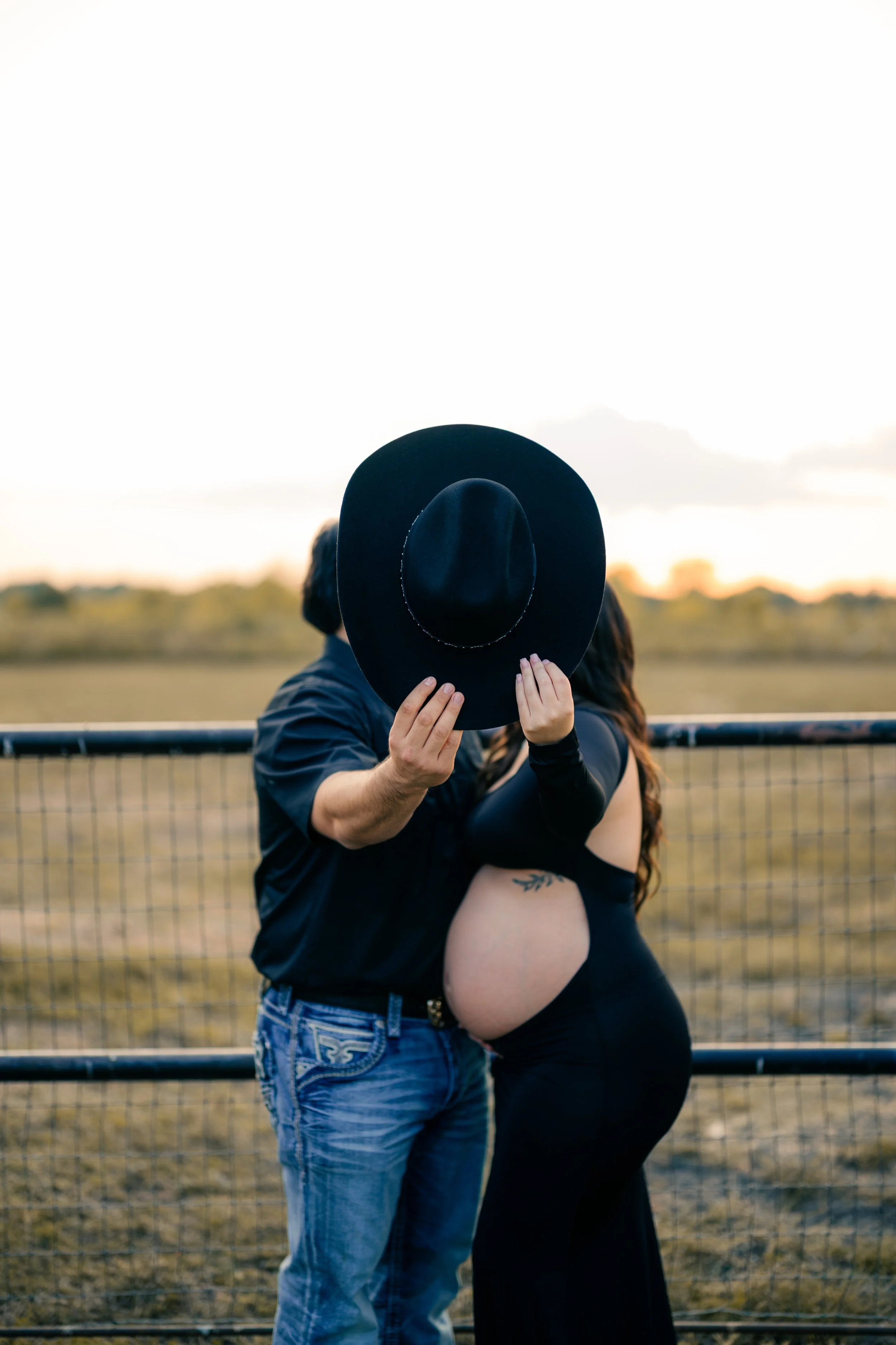 A couple outdoors behind a fence, with the woman showing a pregnant belly, and the man holding a large black hat in front of their faces.