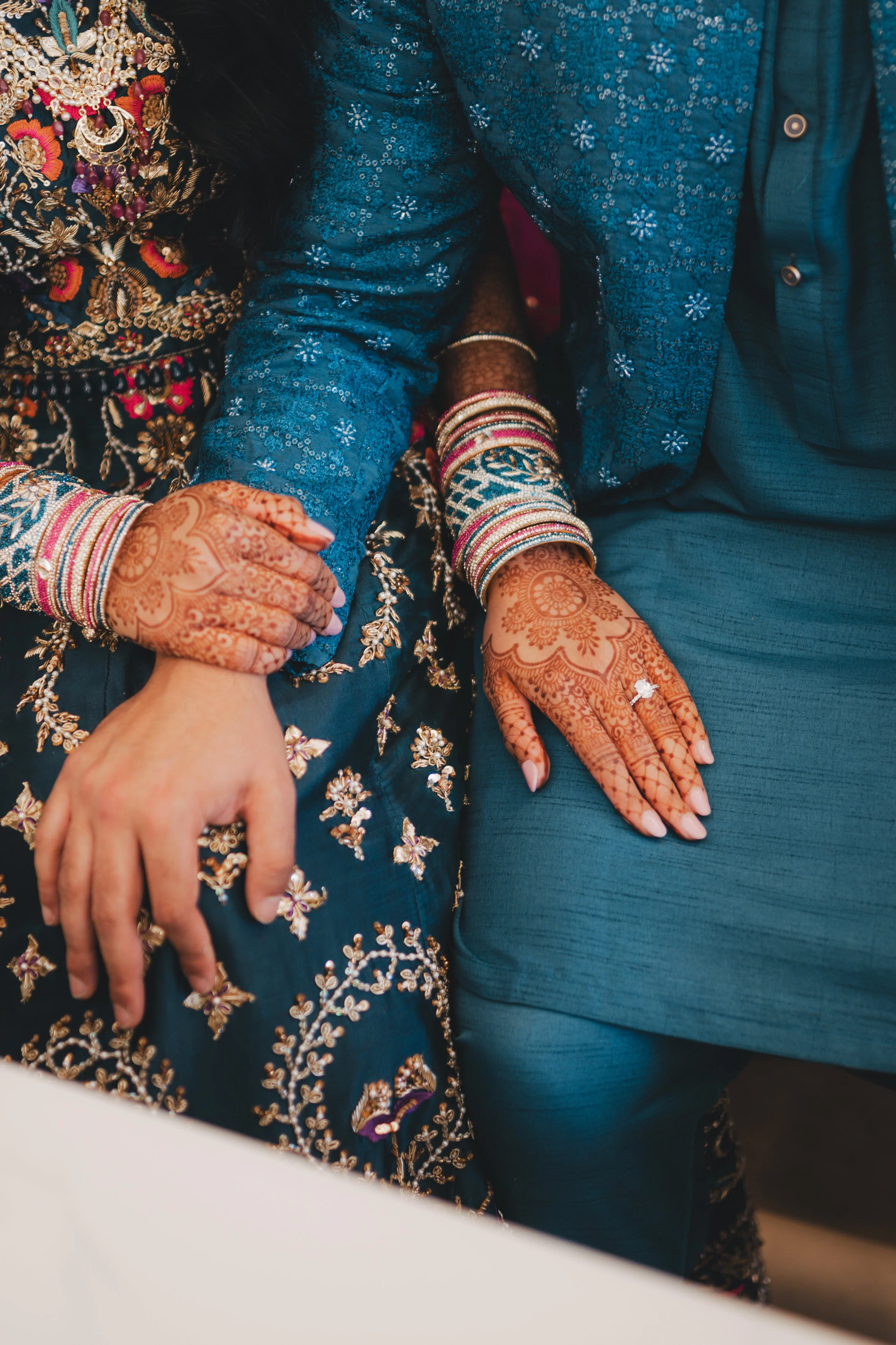 Close-up of a bride and groom's hands, showing intricate henna designs on their hands and adorned with colorful bangles. The bride is wearing a heavily embroidered dark blue dress, and the groom is dressed in a blue outfit with floral embellishments.