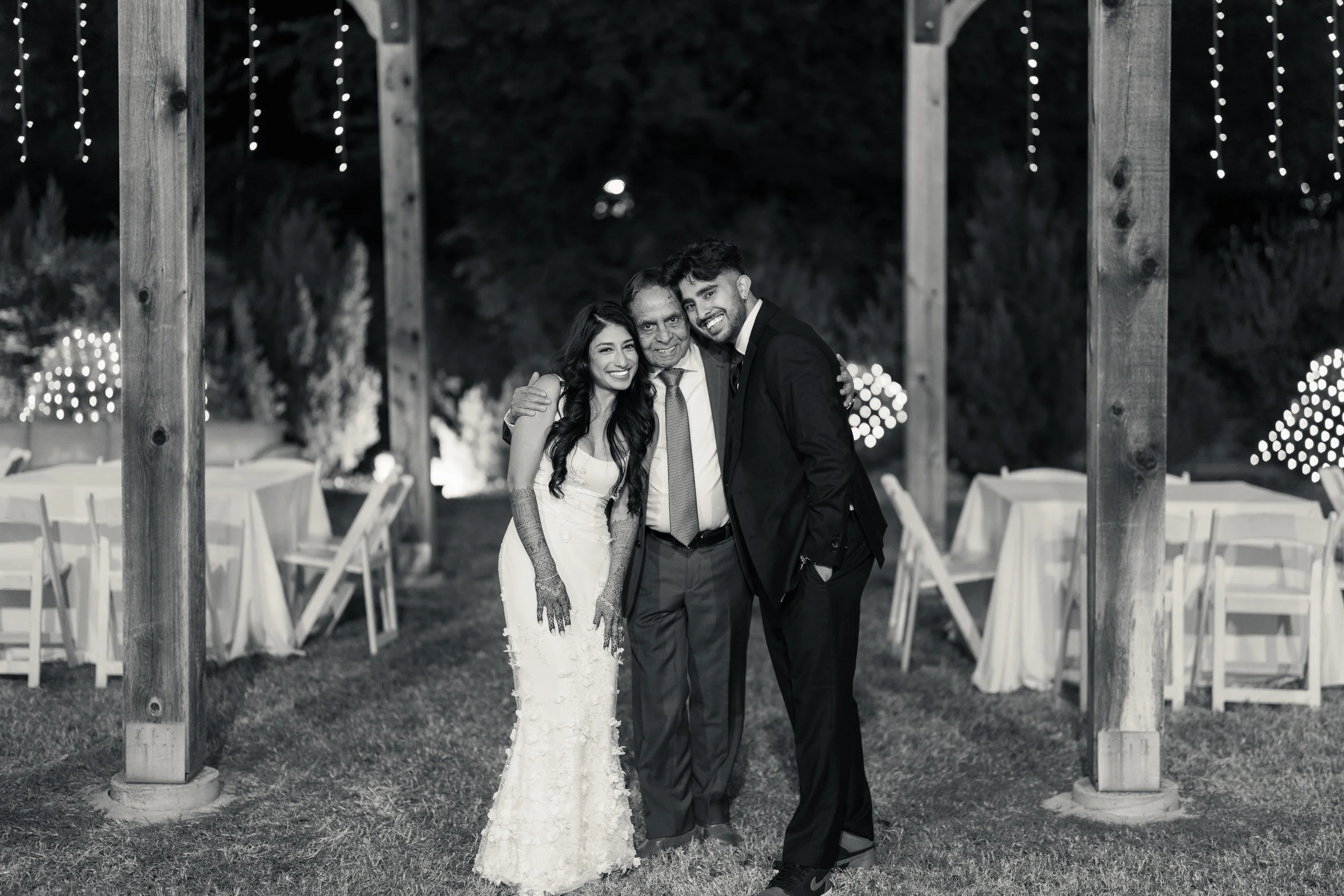 A black and white photo of three people celebrating at a wedding reception, with two men and a woman, smiling and hugging. The woman is wearing a wedding gown, and the man on the right is dressed in a suit. They are standing outdoors under a wooden s