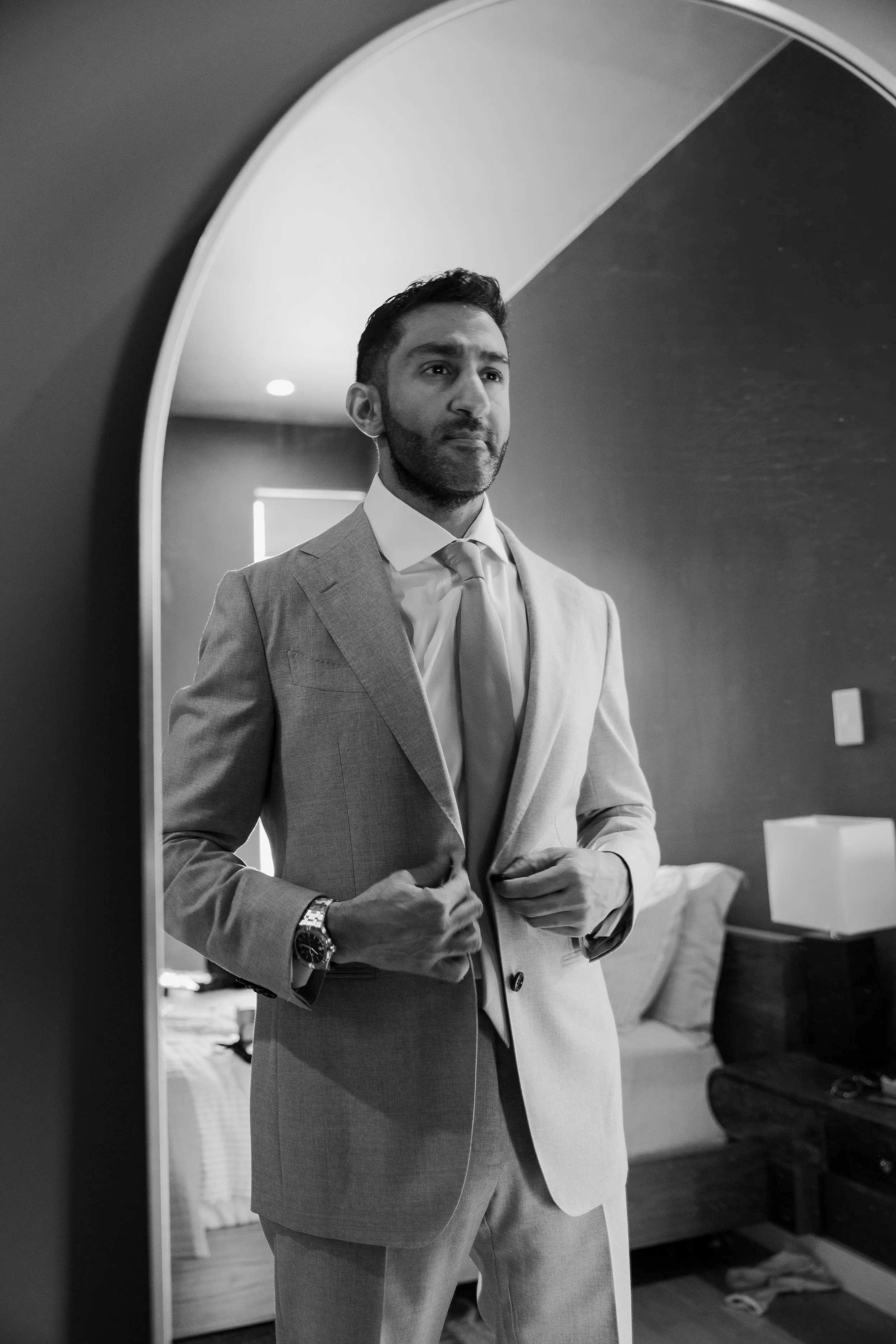 A man in a light-colored suit and tie standing before a mirror, adjusting his suit jacket, in a bedroom with a bed and window in the background.