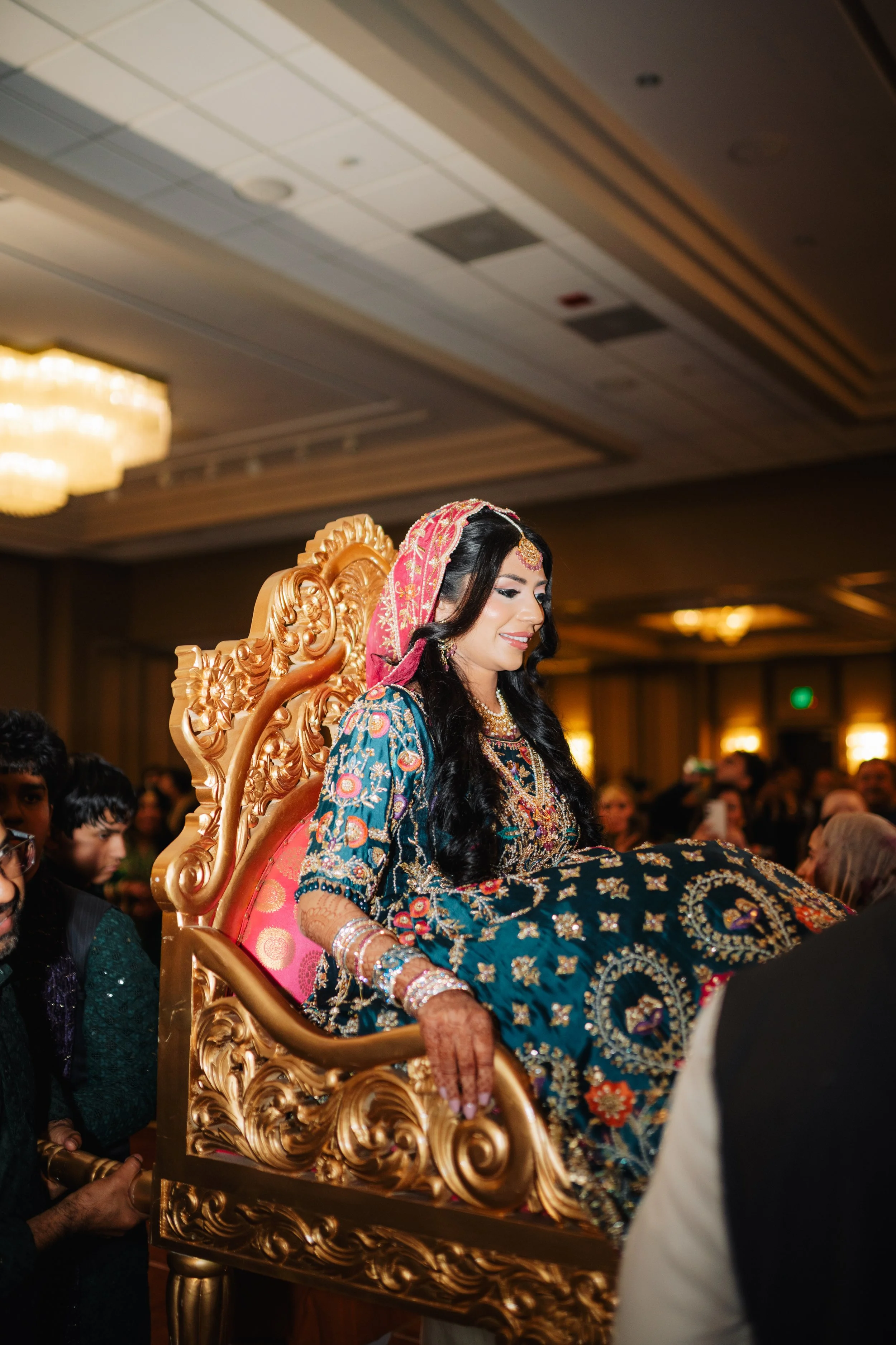A woman dressed in traditional South Asian attire, adorned with jewelry and a pink head covering, is sitting on an ornate golden throne during a celebration or wedding ceremony, surrounded by a crowd in an indoor venue.