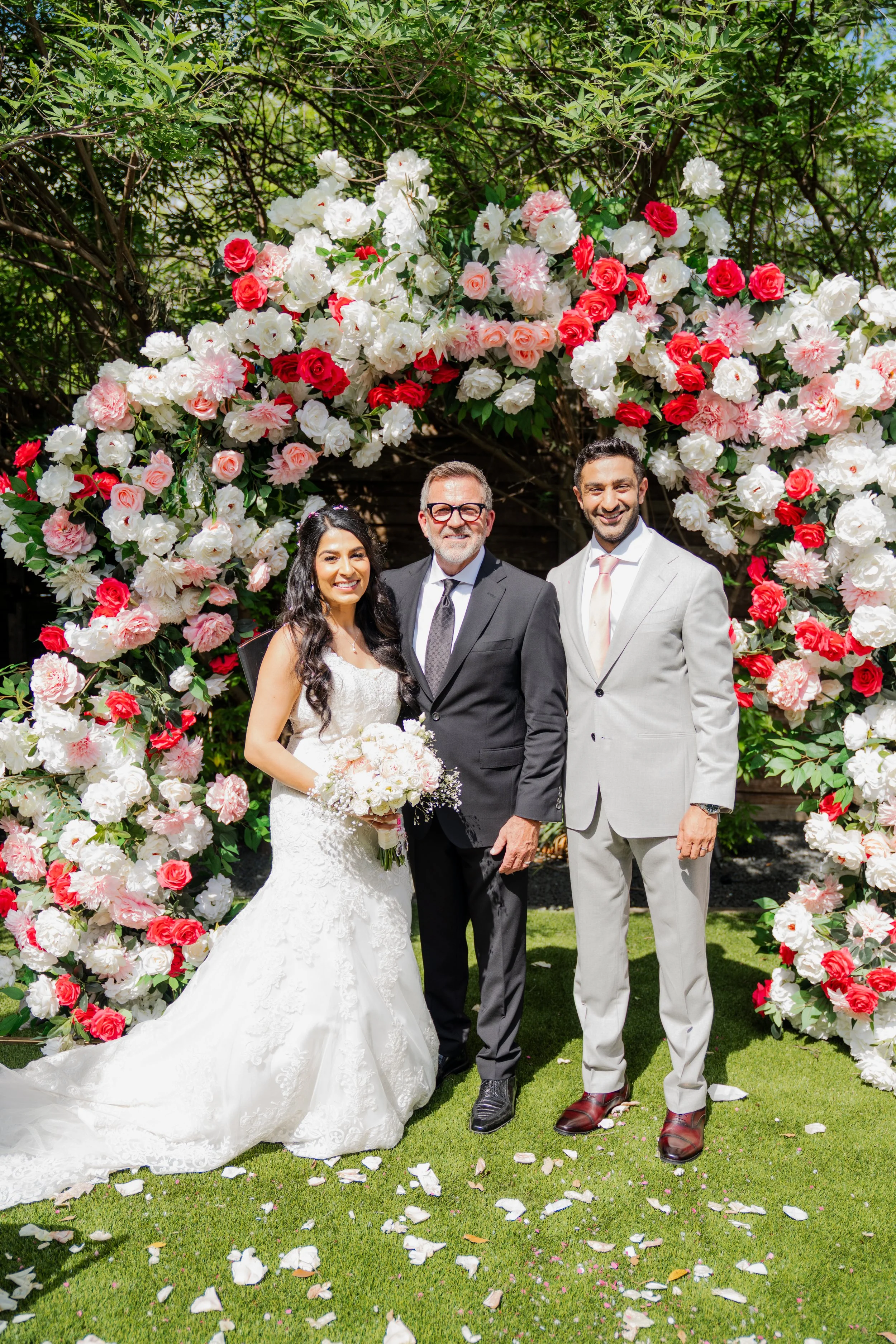 A wedding ceremony with a bride, an older man, and a younger man standing in front of a floral archway made of pink, red, and white flowers. The bride wears a white lace wedding gown, the older man in a dark suit, and the younger man in a light-color