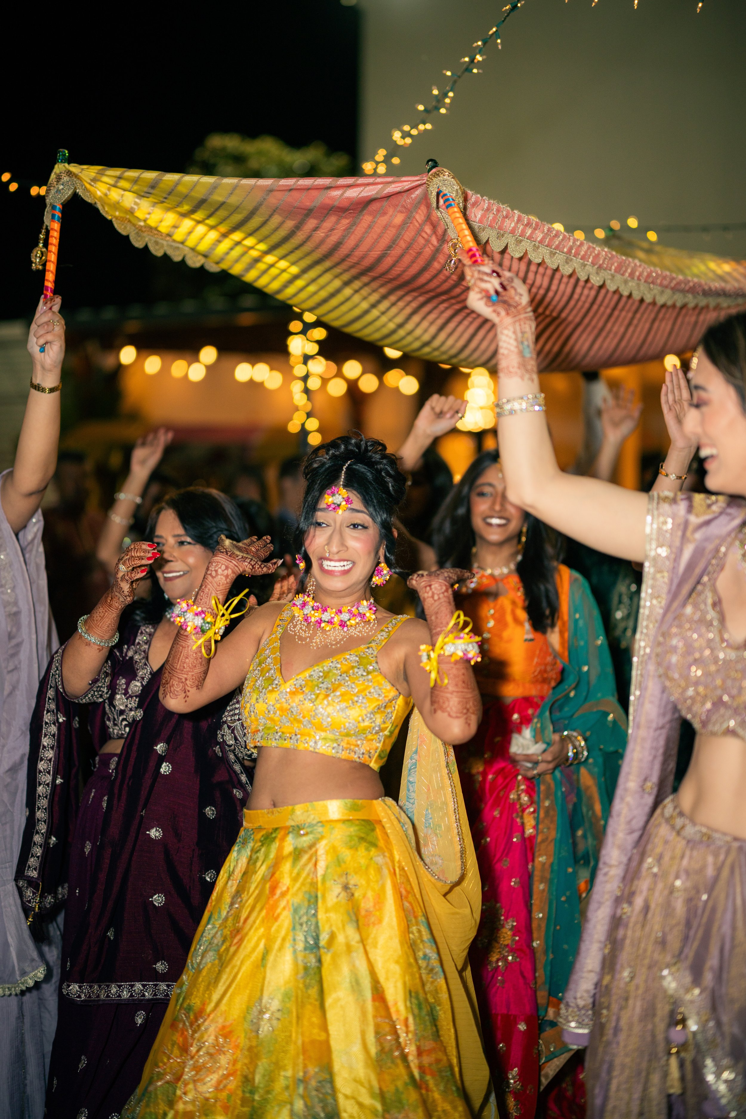 A woman in a yellow traditional outfit is celebrating at a cultural event, surrounded by other women in colorful dresses, under string lights, with a decorated canopy above.