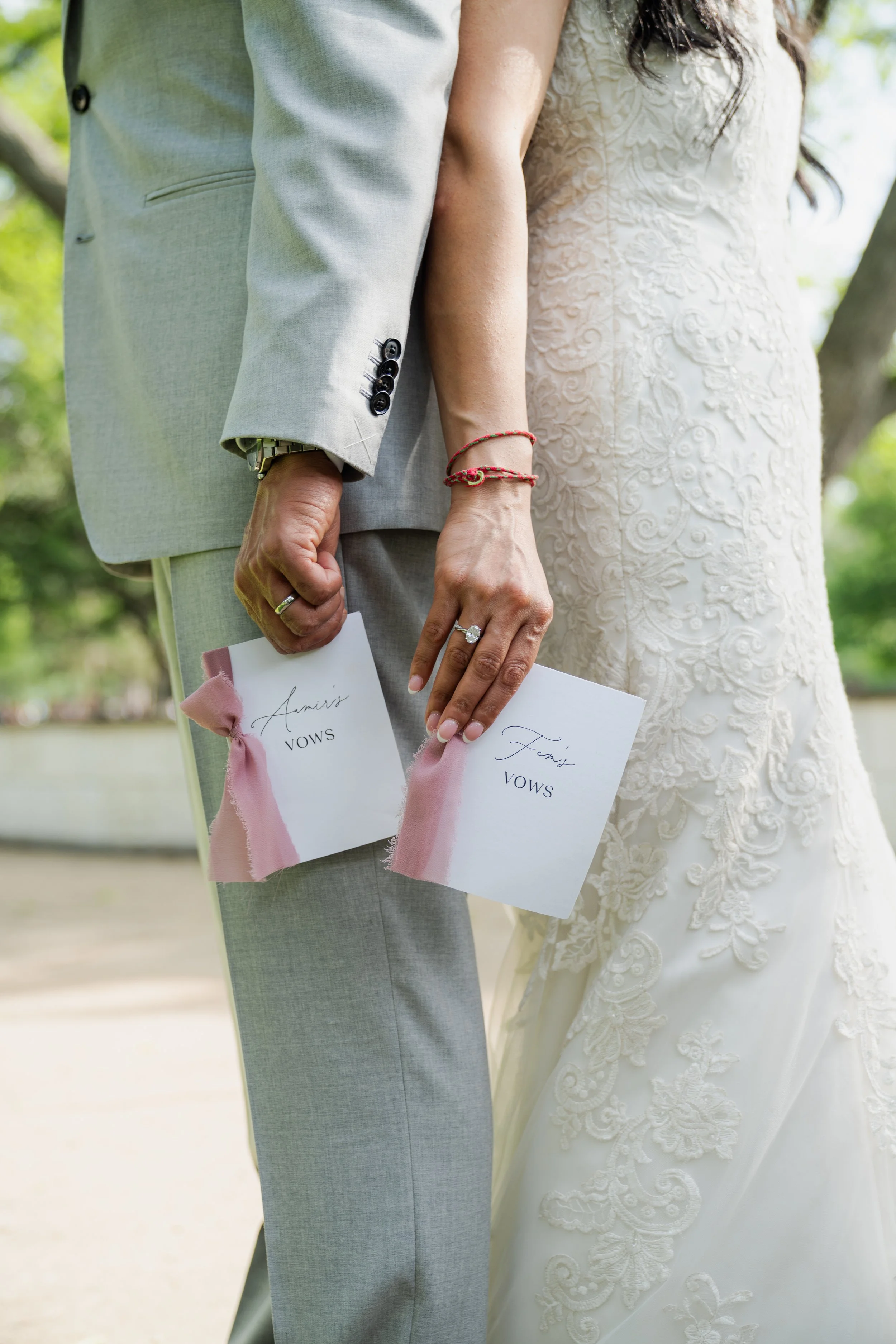 Close-up of a bride and groom holding hands, holding vows with pink ribbons, outdoors with trees in the background.