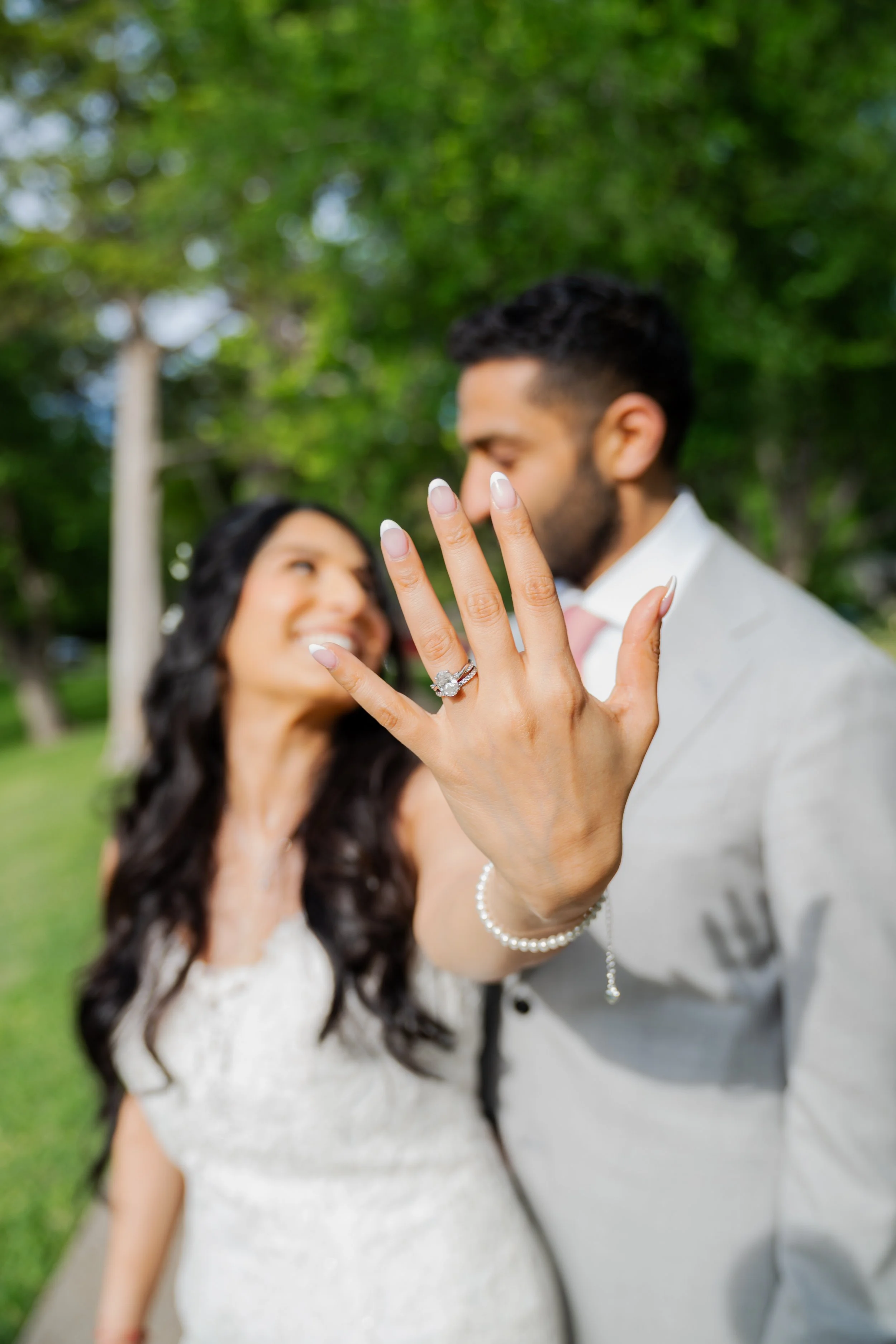A couple standing outdoors in a park, with the woman showing her diamond engagement ring on her finger as the man holds her hand, both smiling and looking at each other, with trees and greenery in the background.