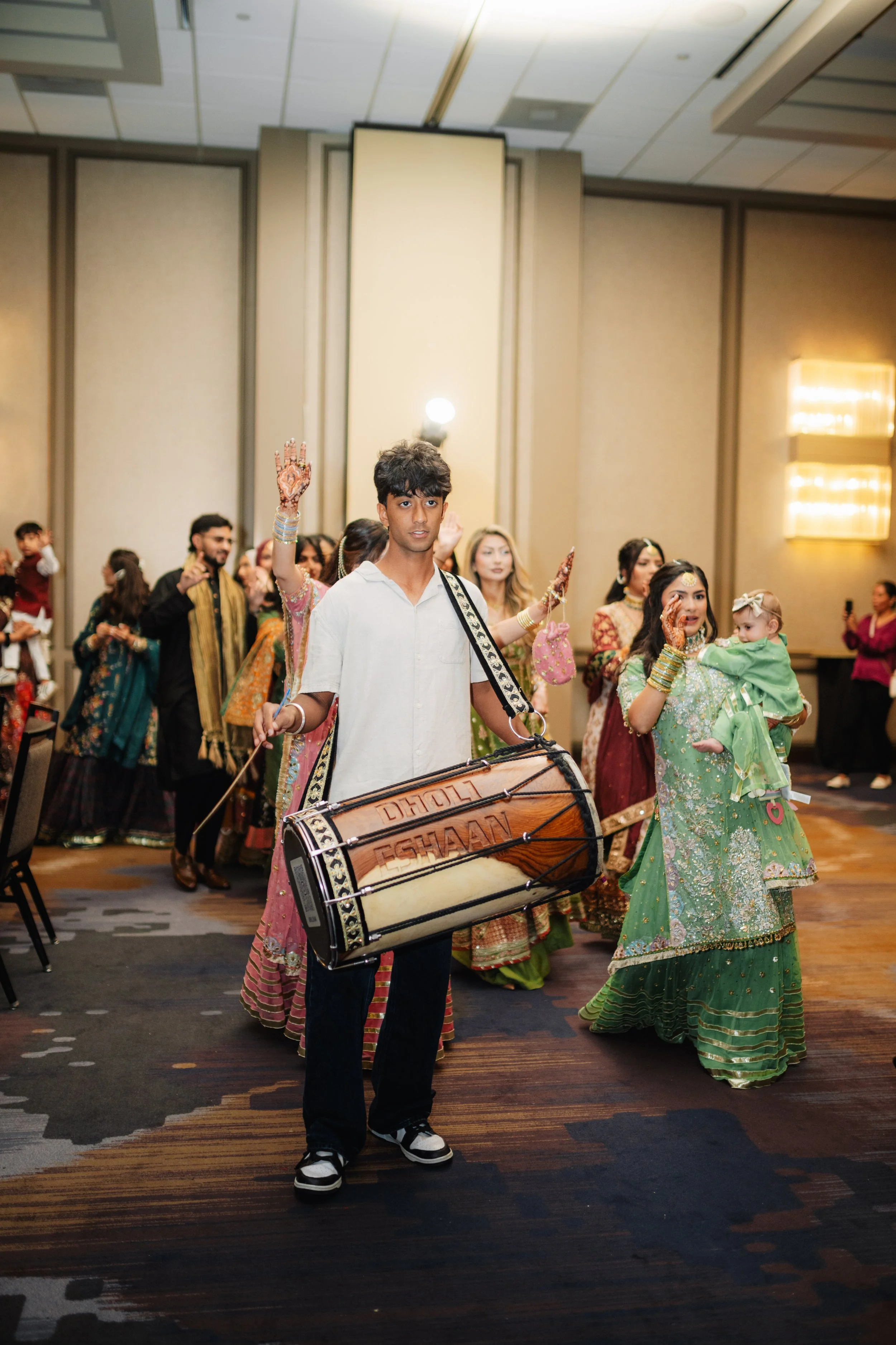 A young man playing a large traditional drum at a cultural celebration with women and children in colorful traditional Indian attire in the background.