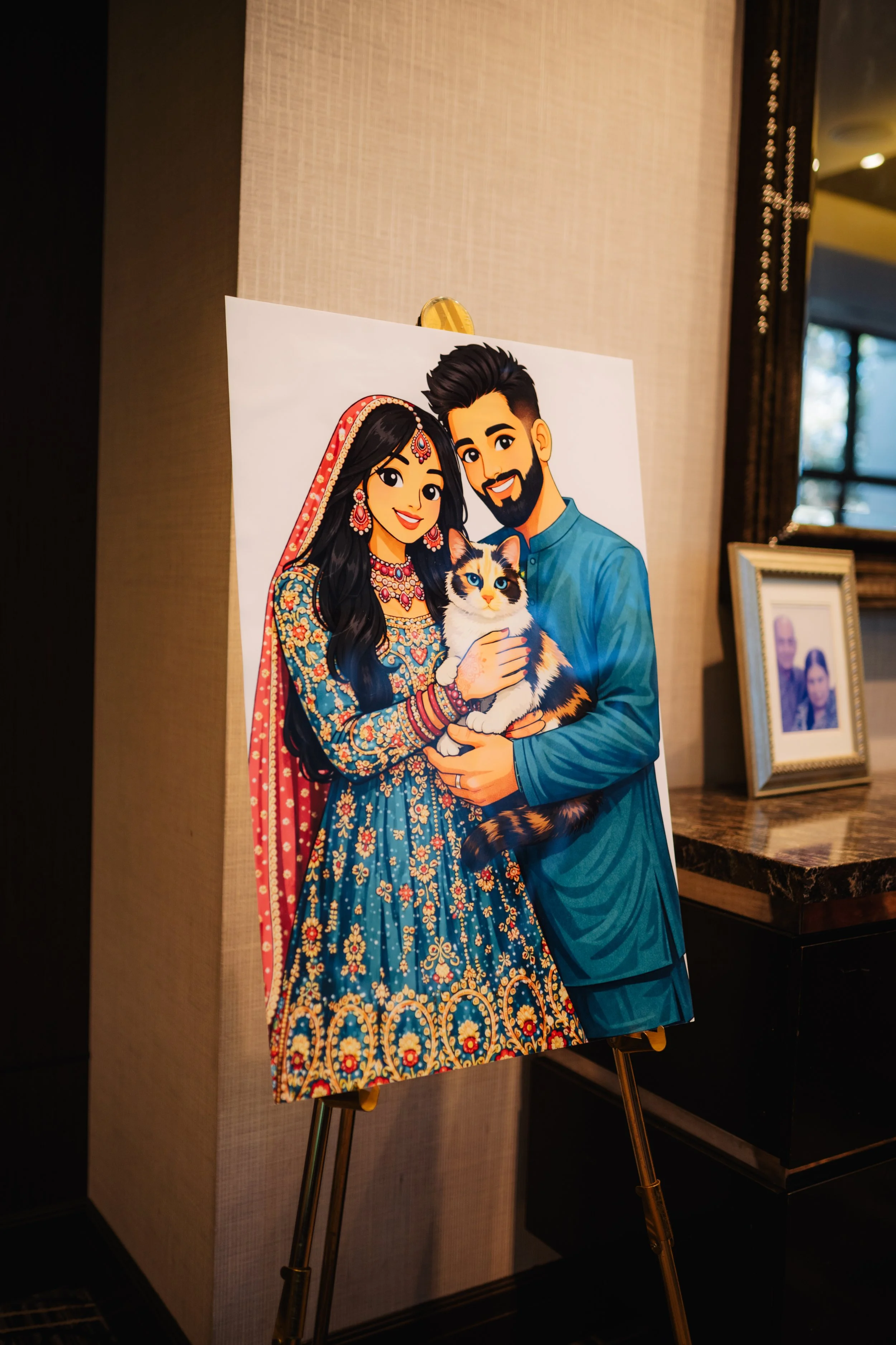 Cartoon poster of a couple dressed in traditional Indian wedding attire holding a calico cat, set on an easel in a hotel lobby.