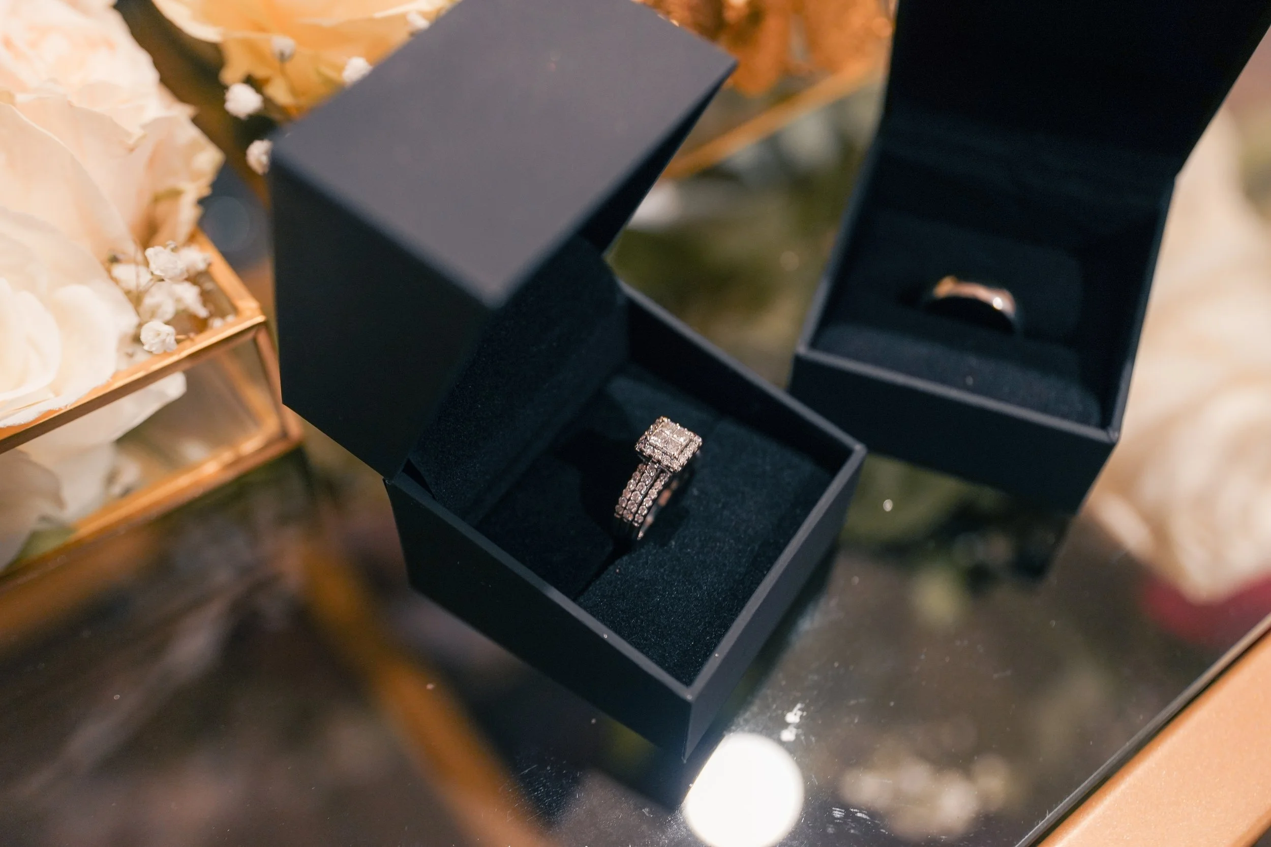 A black jewelry box containing a diamond wedding ring, and another black ring box with a gold band, displayed on a glass table surrounded by white flowers.