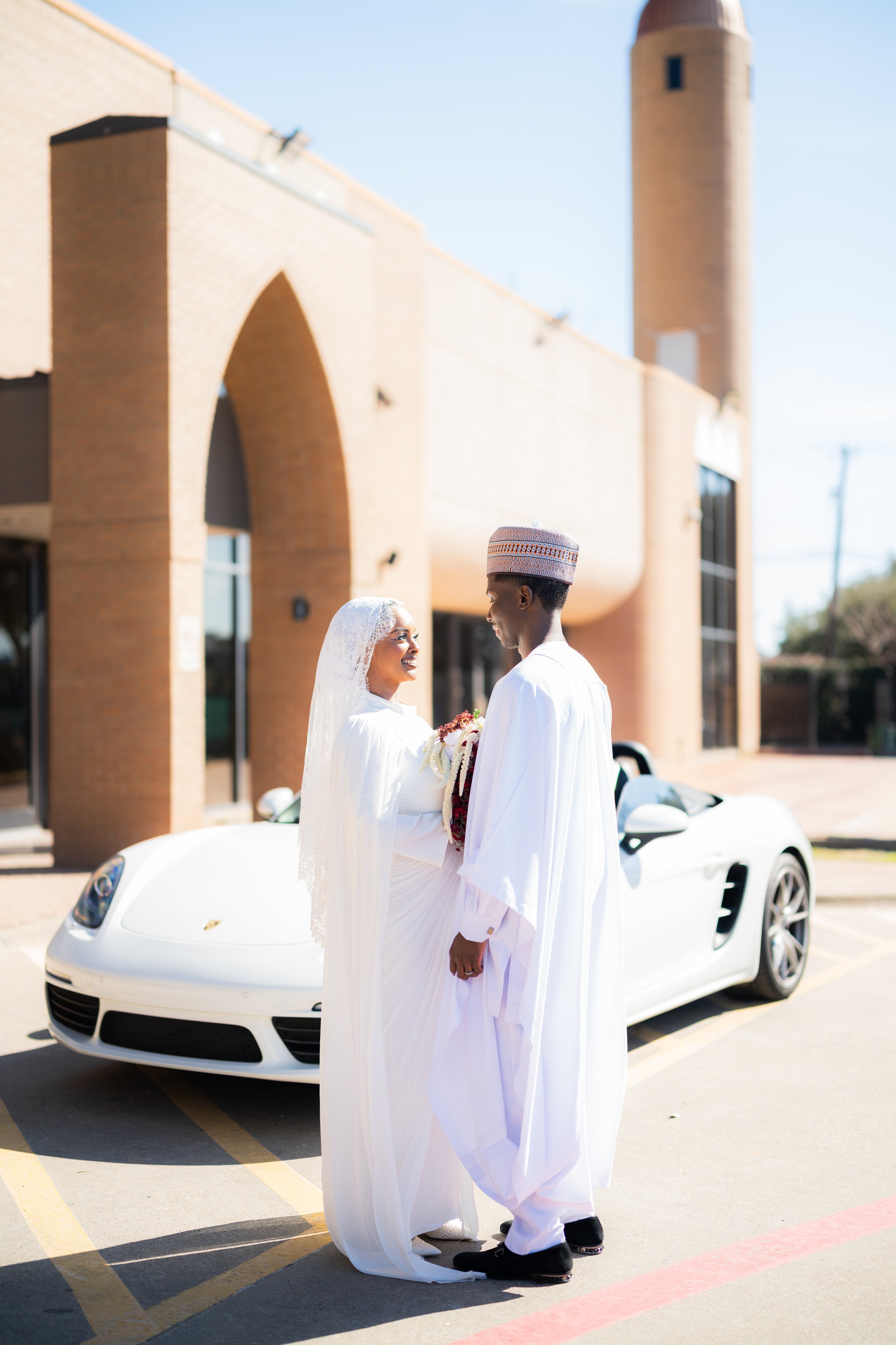 A bride and groom in traditional white wedding attire are smiling and looking at each other while standing in front of a white sports car. The bride is wearing a veil and holding a bouquet, and the groom is wearing a hat. They are outdoors in a parki