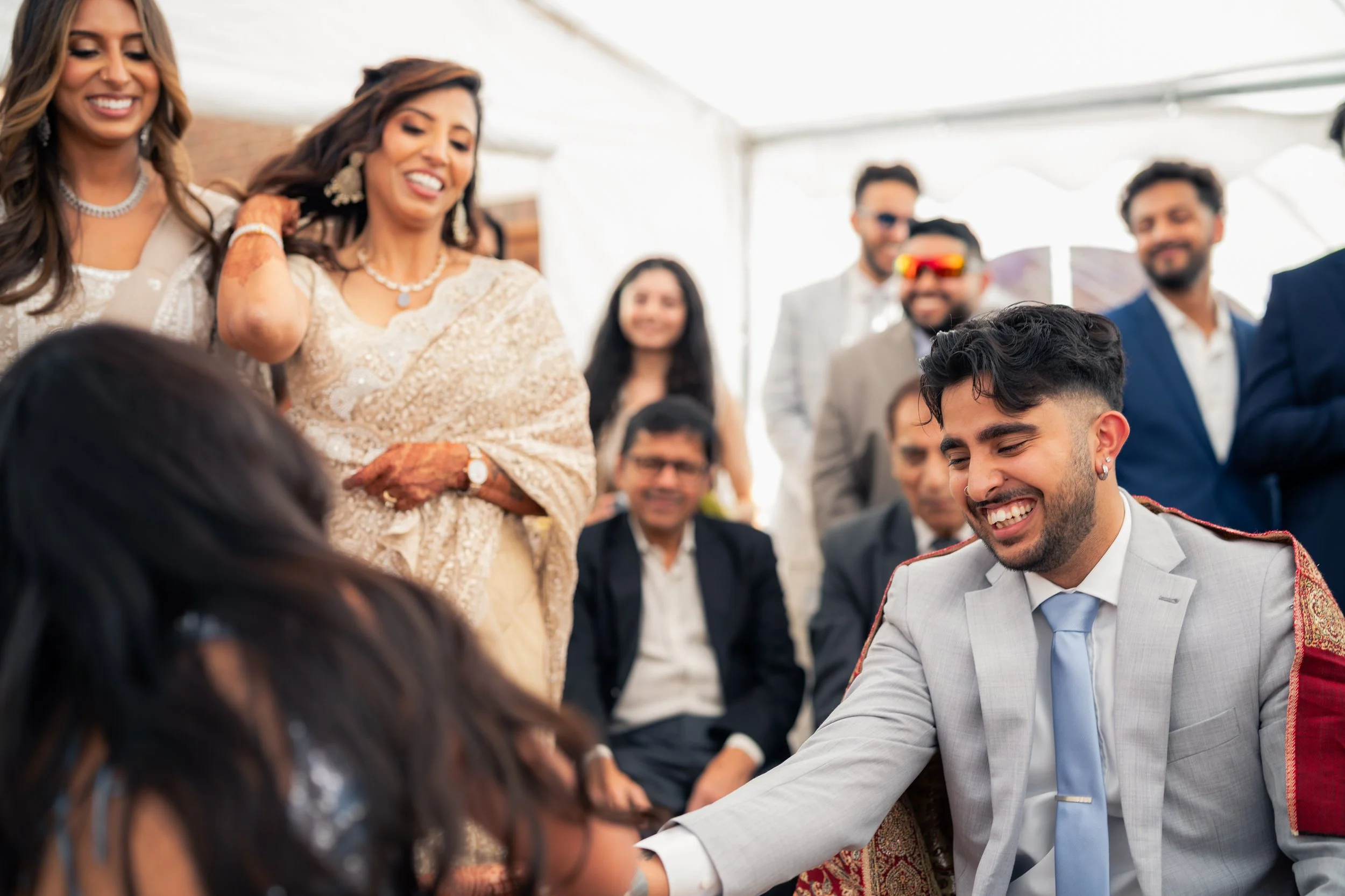A group of people at a celebration, with a man in a gray suit and blue tie smiling and shaking hands with a woman whose face is not visible. The group is dressed in formal attire, and some women are wearing traditional South Asian clothing. Everyone 