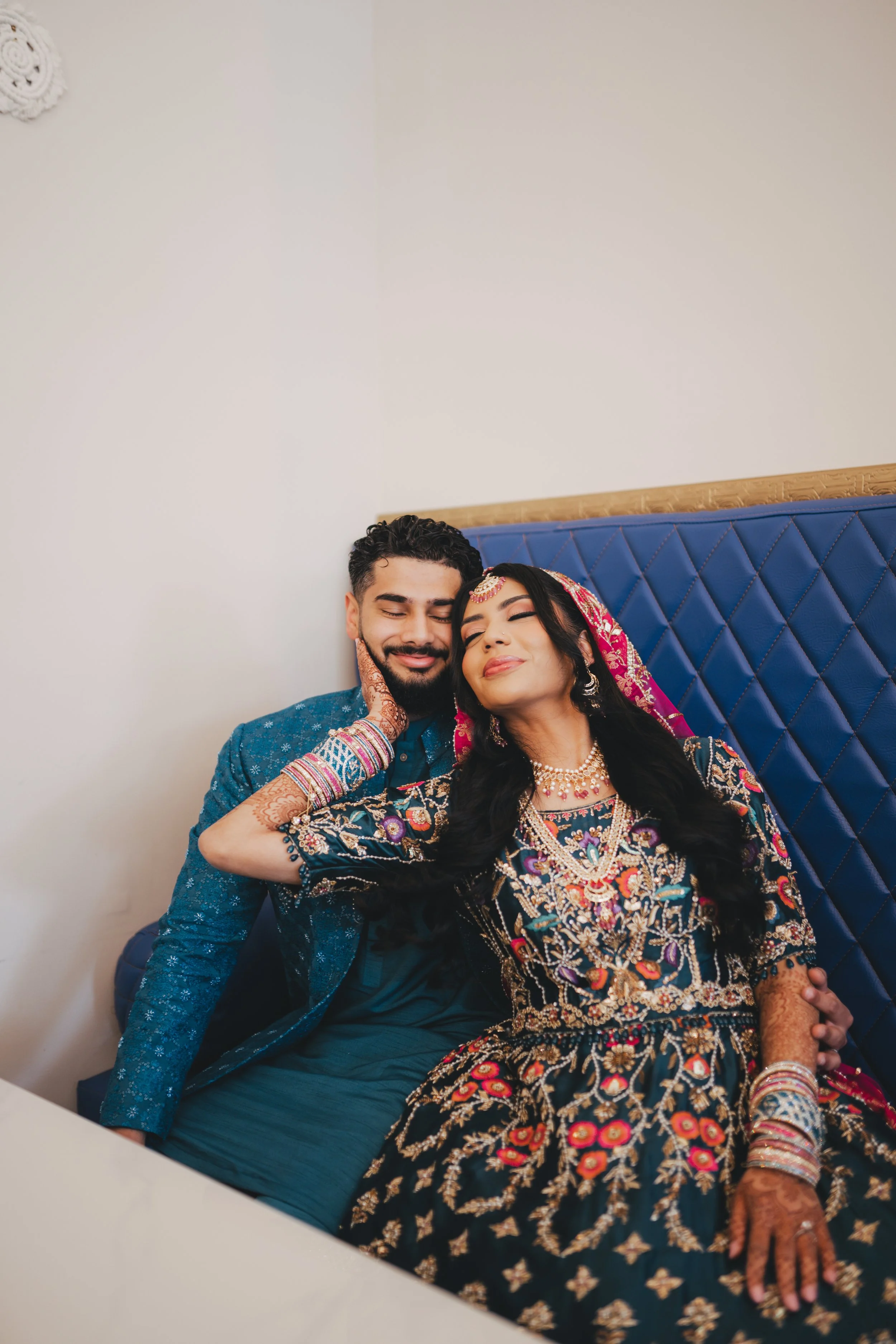 A couple dressed in traditional Indian wedding attire sitting close together on a blue quilted headboard, smiling with eyes closed, in a room with plain wall decor.