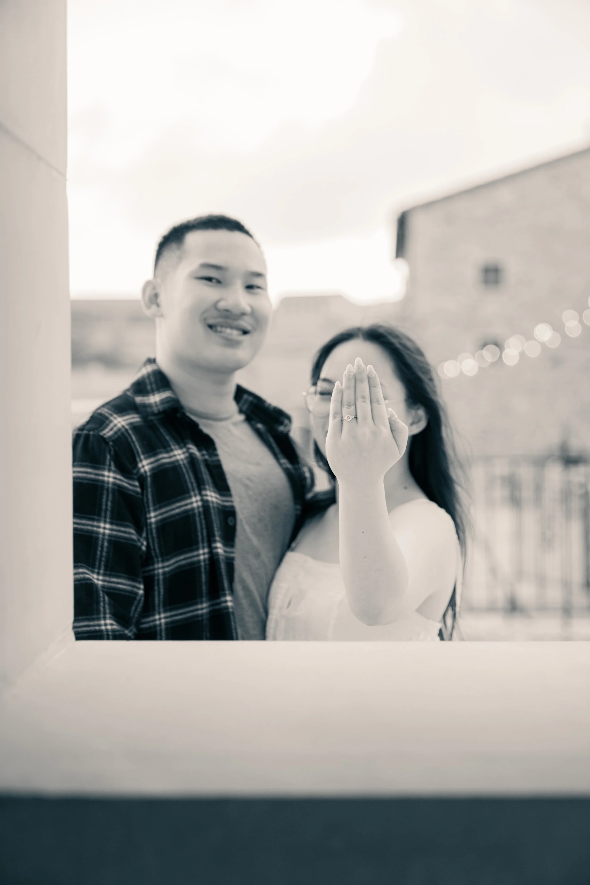 A couple is standing outside, with the woman showing her ring on her finger. The photo is in black and white, and the woman is partly covering her face with her hand to display the ring.