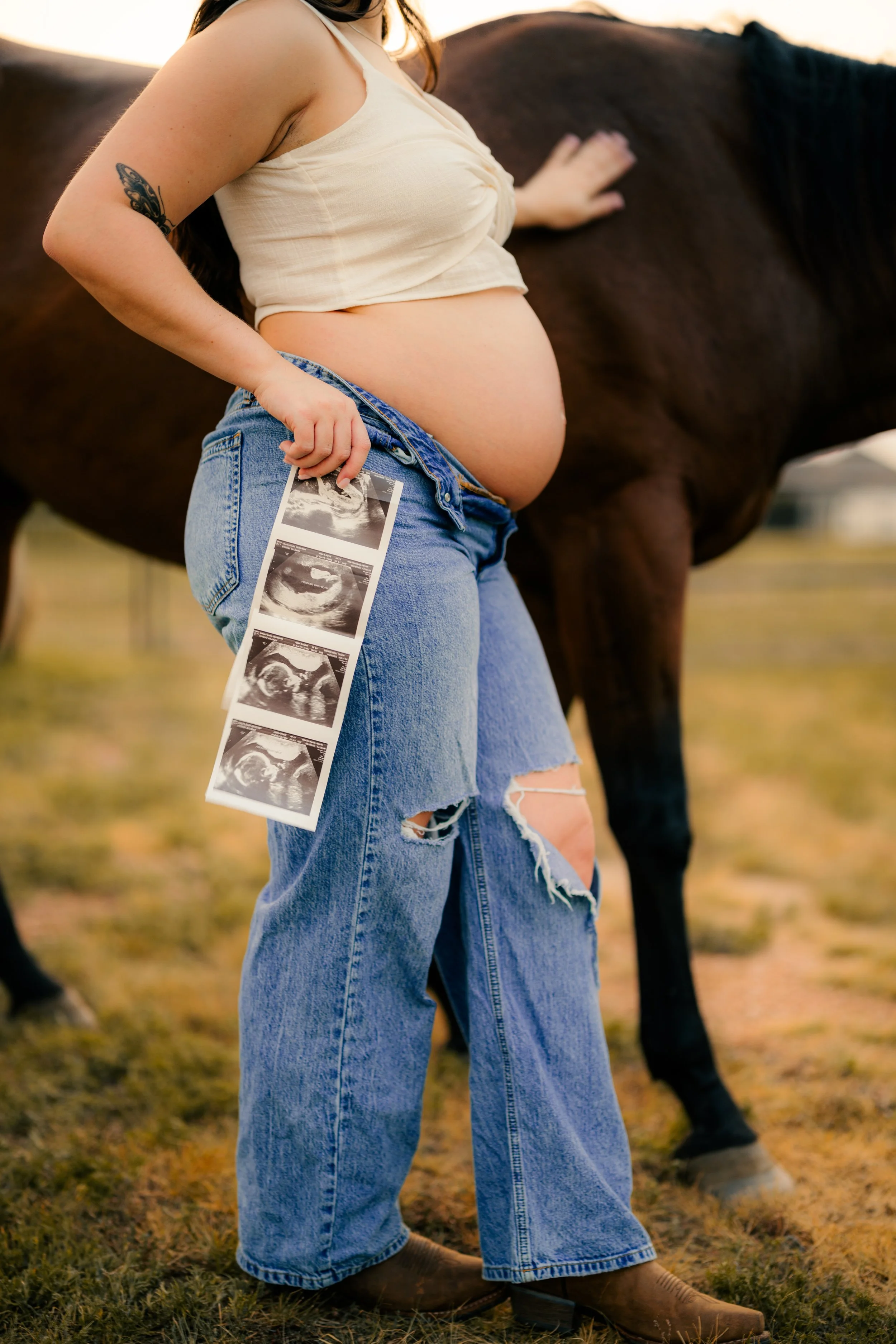 A pregnant woman holding ultrasound images, standing outdoors with a horse in the background.