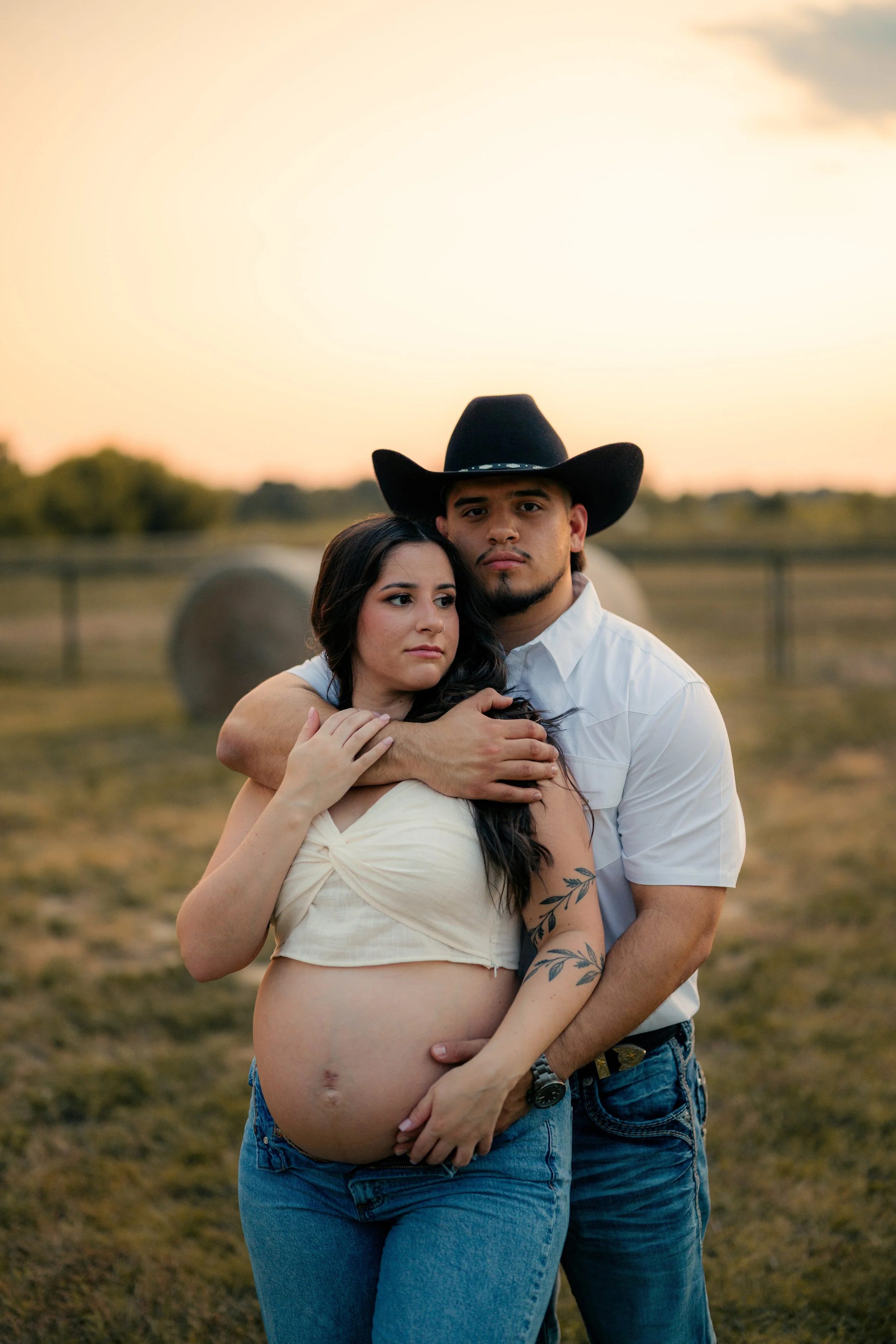 A pregnant woman and a man, possibly her partner, standing outdoors in a field at sunset. The woman has long dark hair, and the man is wearing a cowboy hat and white shirt. They are embracing, with the man’s arm around the woman’s shoulders and her h