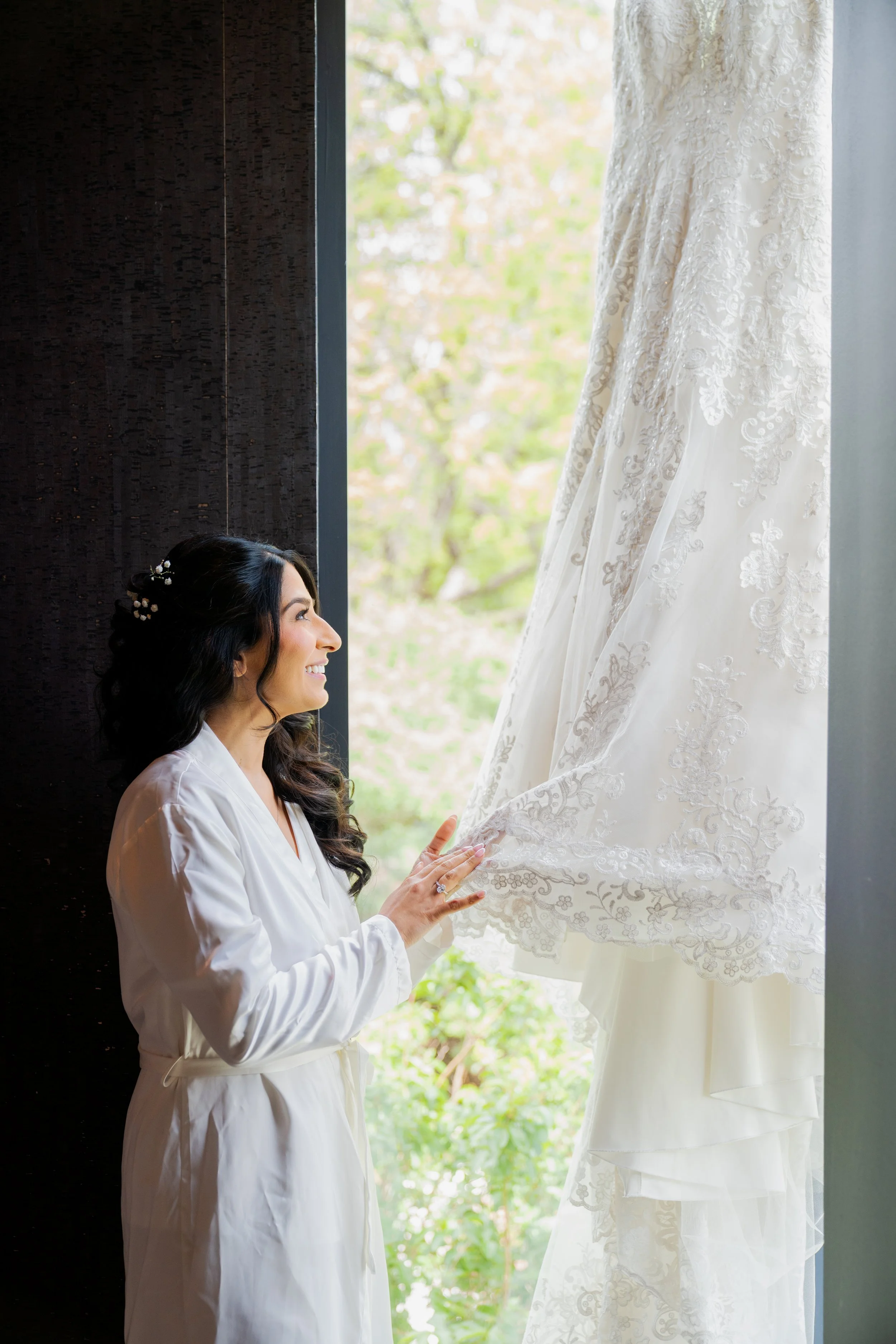 A woman in a white robe smiling while looking at a wedding dress hanging near a window, with trees visible outside.