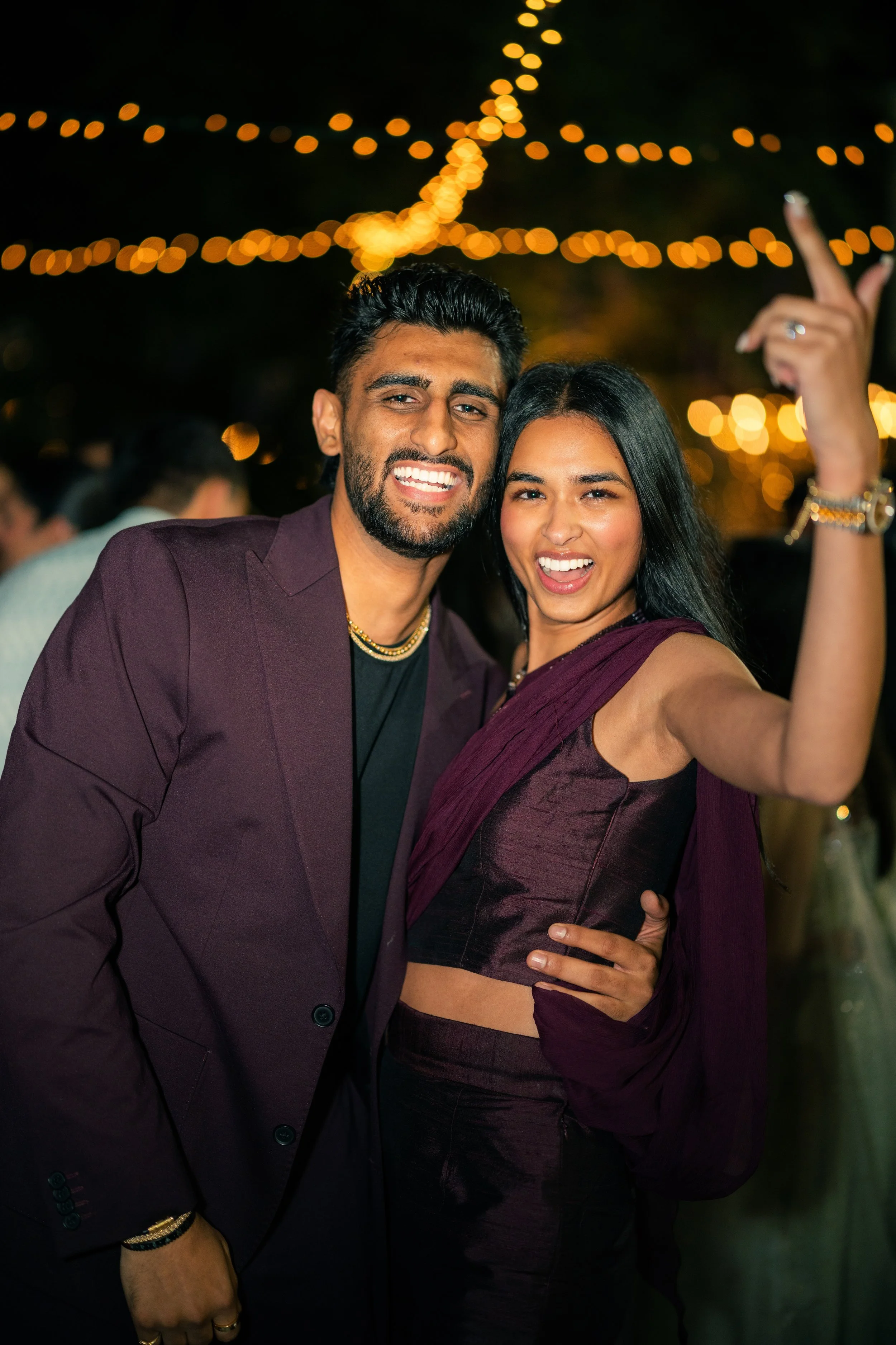 A smiling man and woman in stylish dark purple outfits celebrating at a festive outdoor event at night, with string lights in the background.