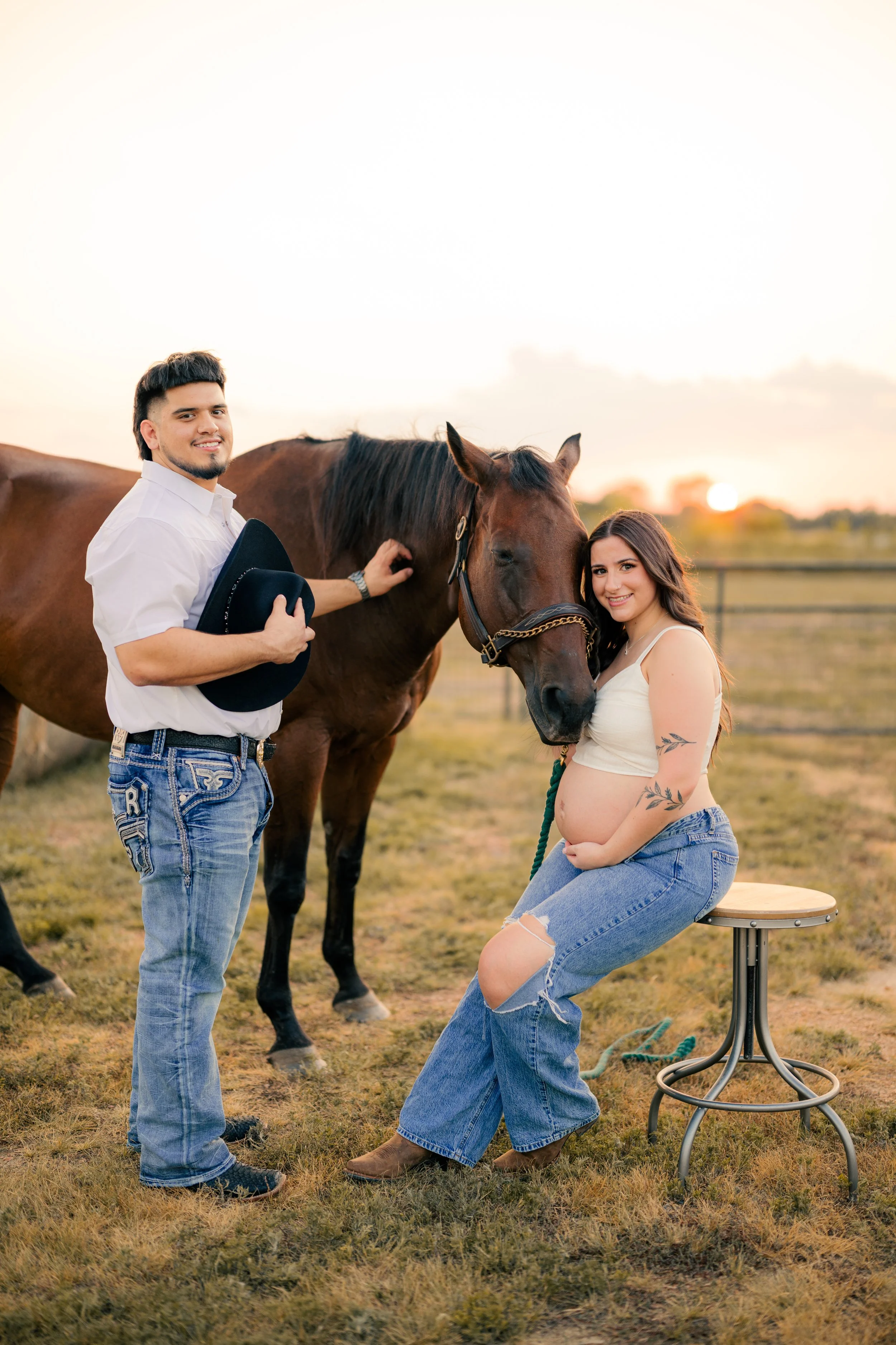 A pregnant woman and a man with a brown horse outdoors at sunset, woman sits on a stool, man stands holding a hat, the woman pets the horse.