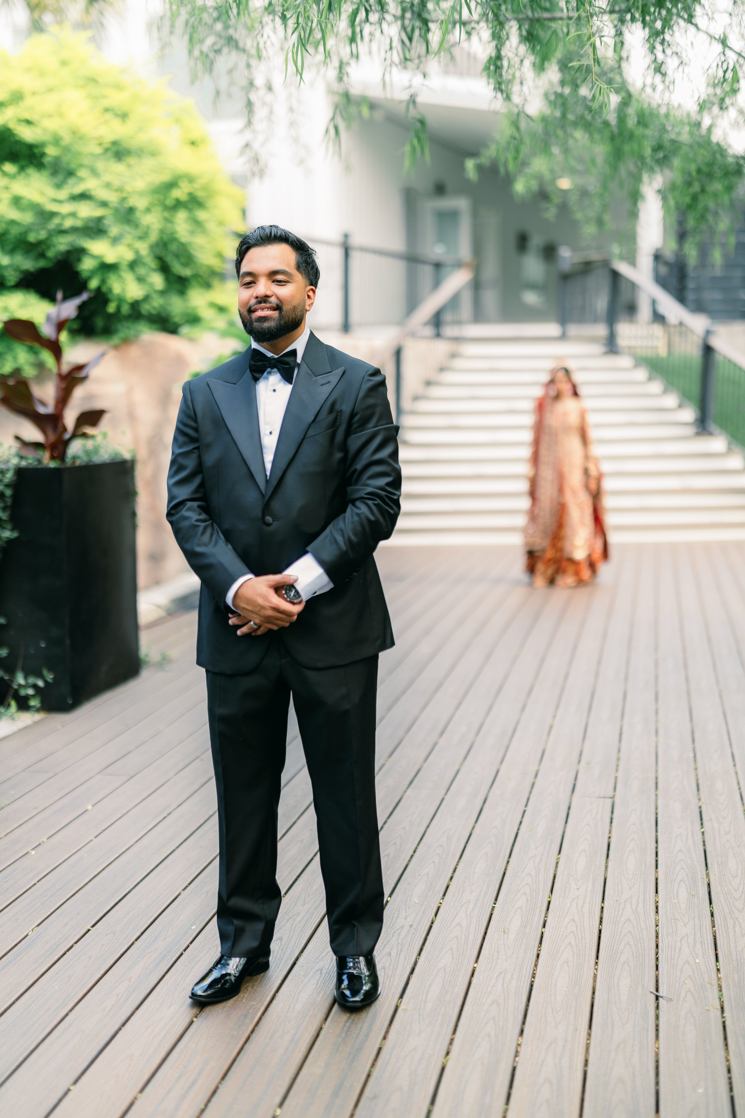 A man in a black tuxedo with a bowtie, standing outdoors on a wooden deck with greenery around, smiling with a woman in a dress and heels in the background on stairs.