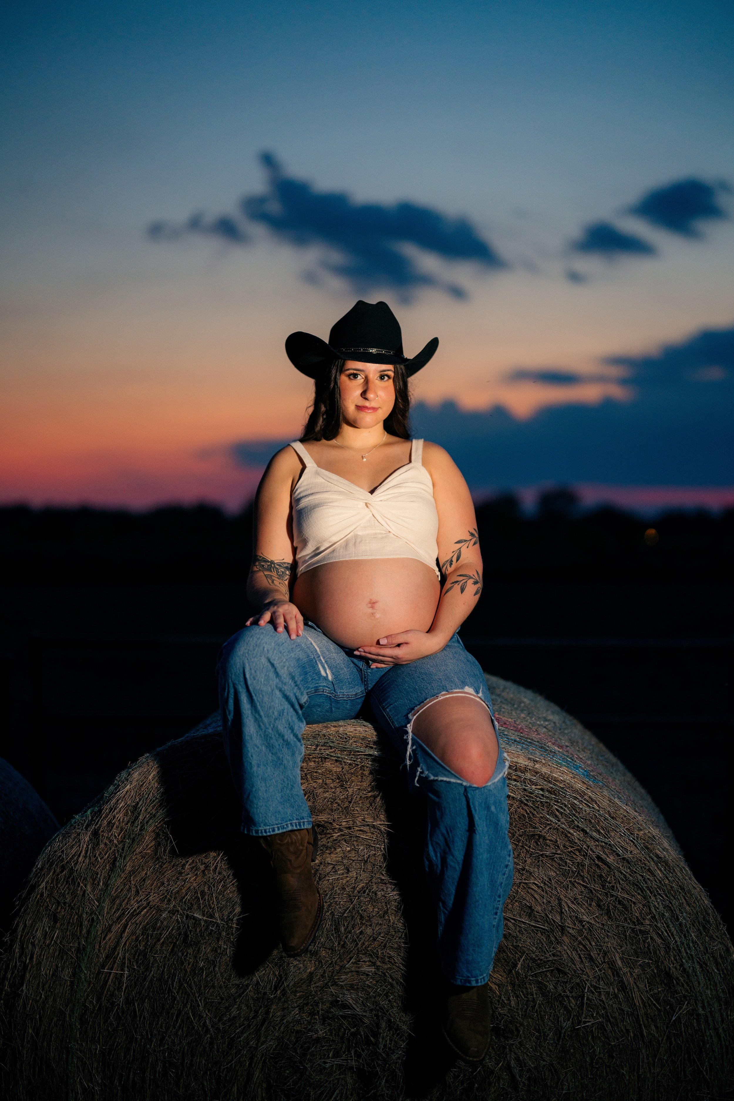 Pregnant woman wearing a cowboy hat, sleeveless top, ripped jeans, and boots sitting on hay bale during sunset.