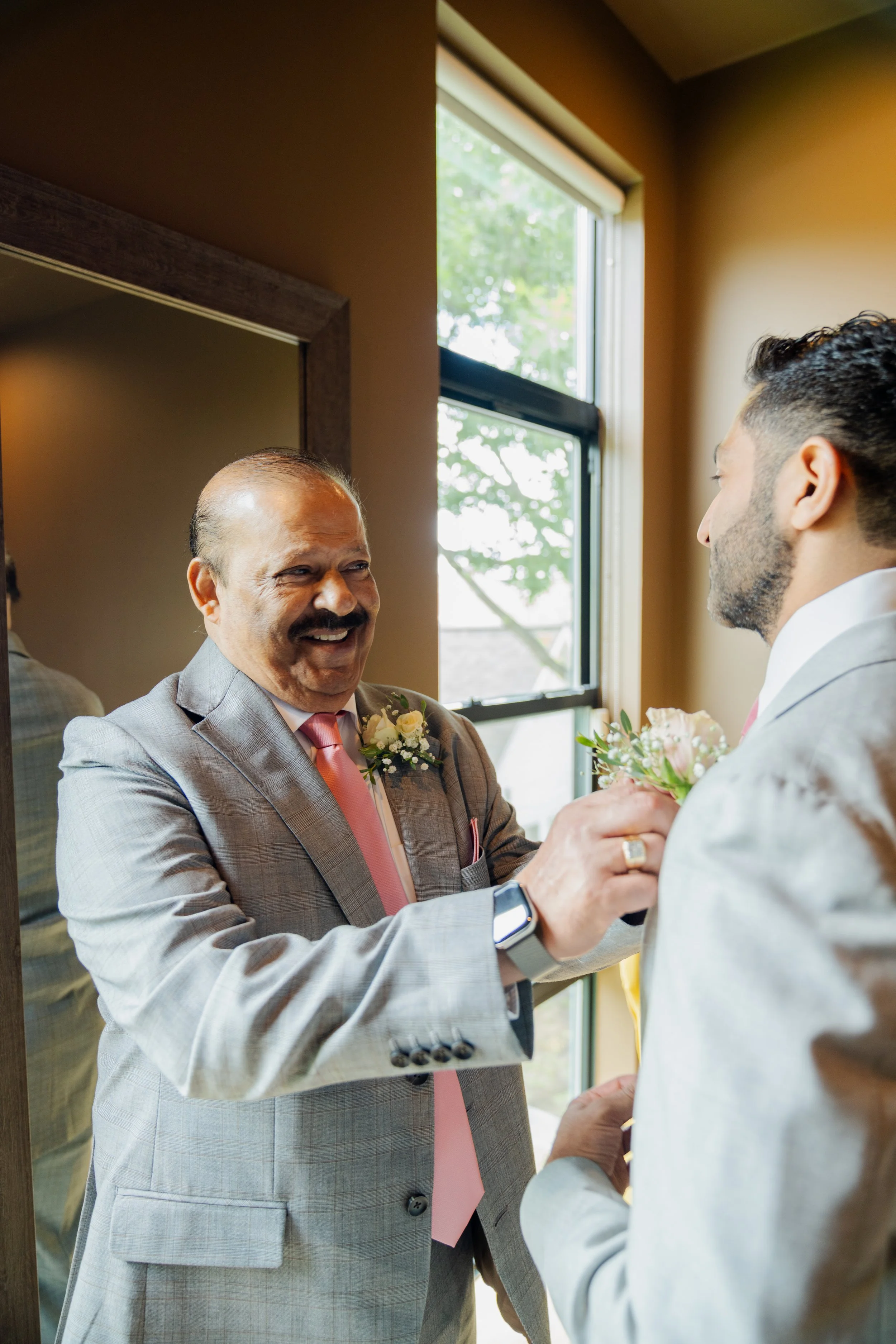 An older man in a gray suit with a pink tie is smiling and holding a pink and white bouquet as he faces a younger man in a white suit, indoors near a window with green trees outside.